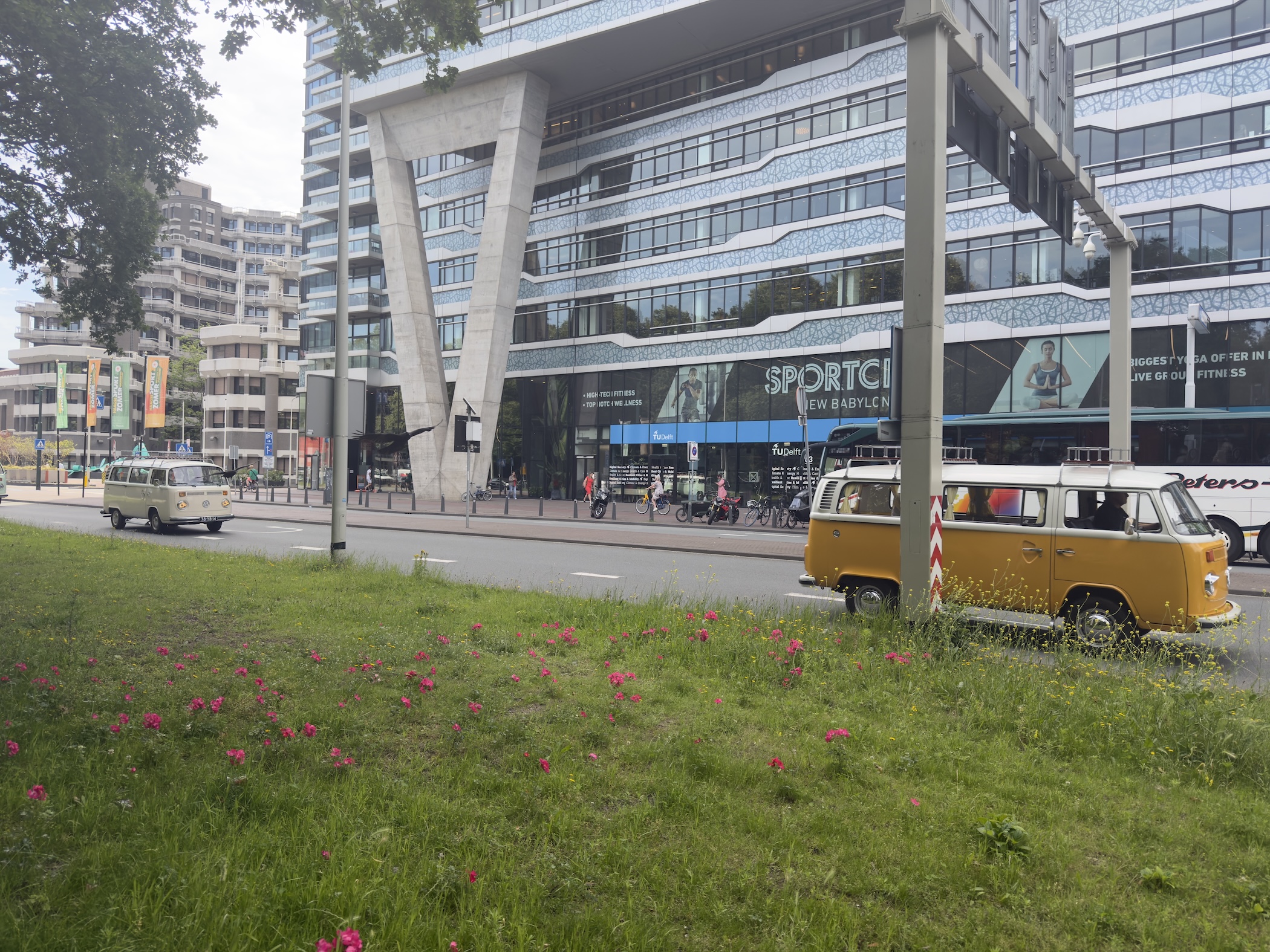 Vintage yellow camper van parked on grass before a modern blue office building, with poppies nearby.