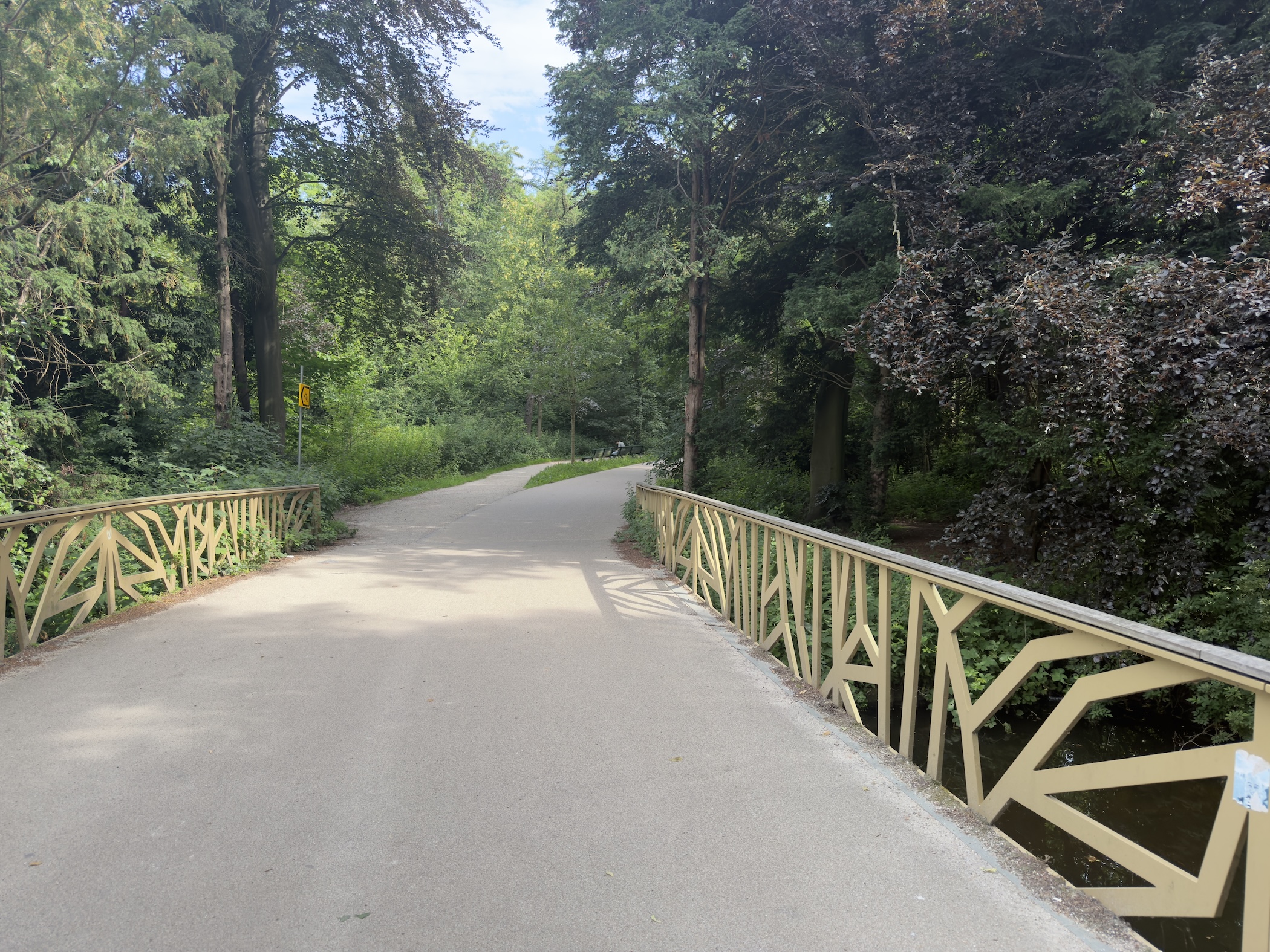 Cycle path crossing an ornate wooden footbridge into dense green woodland.