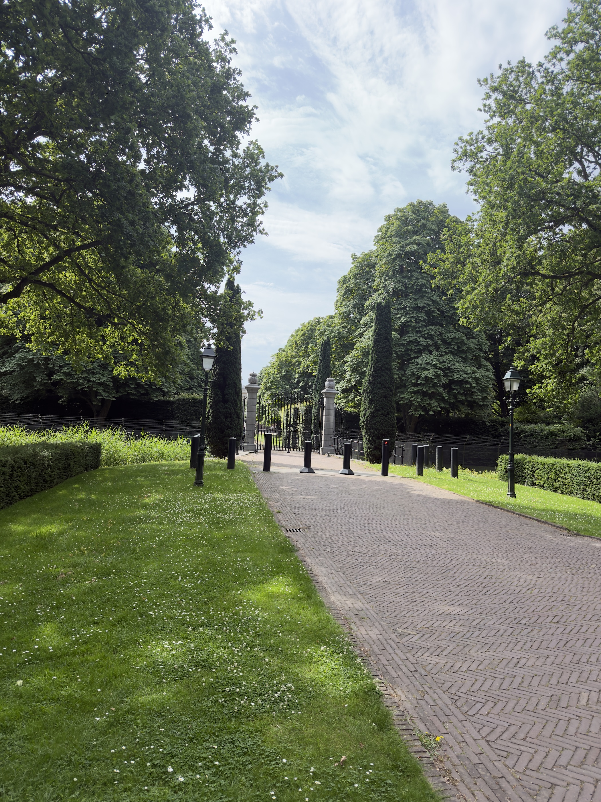 Brick-paved avenue lined with conifers leading to ornamental gateway pillars under a cloudy sky.