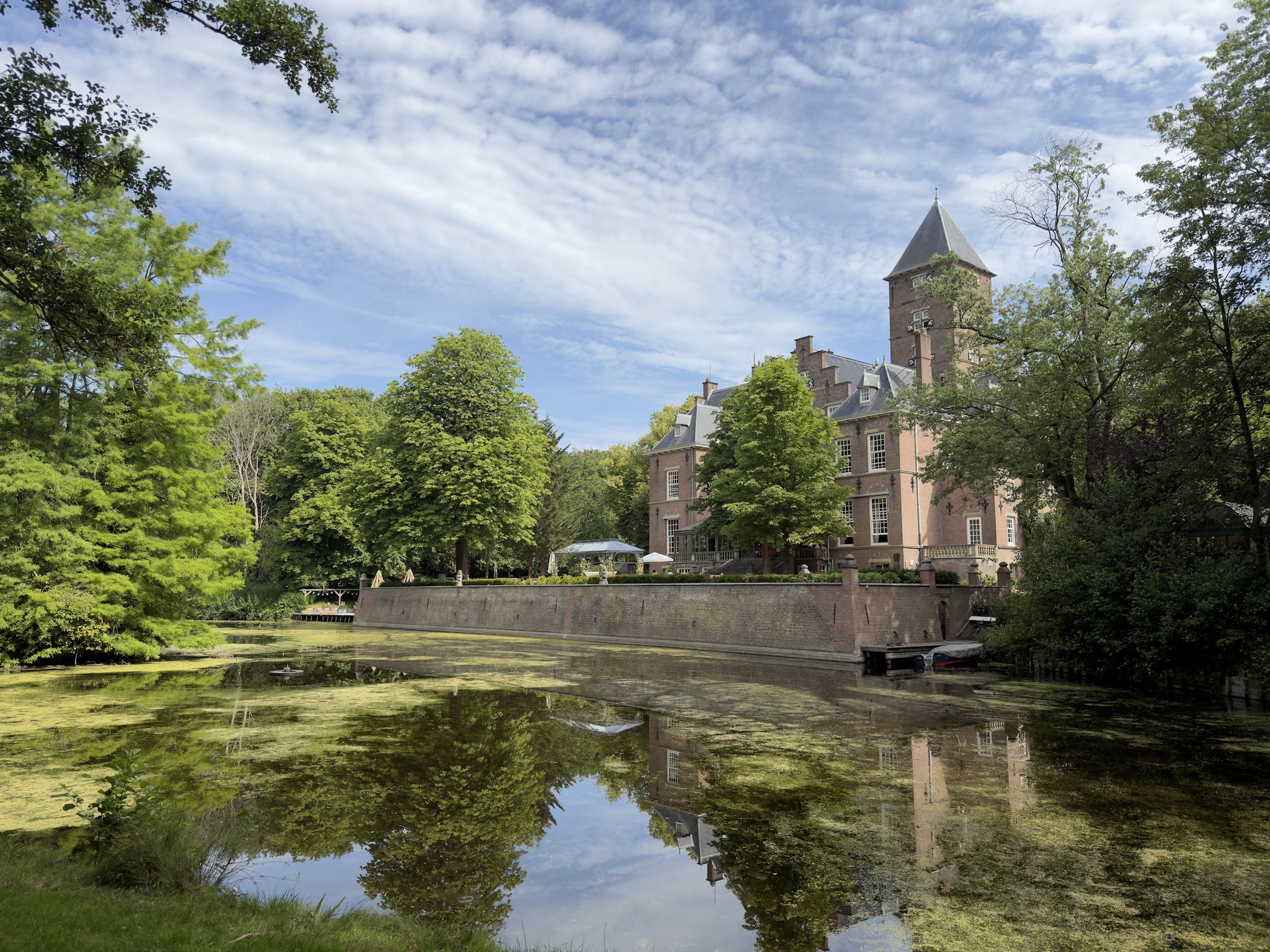 A turreted brick country house reflected in a still moat ringed by trees under blue sky.