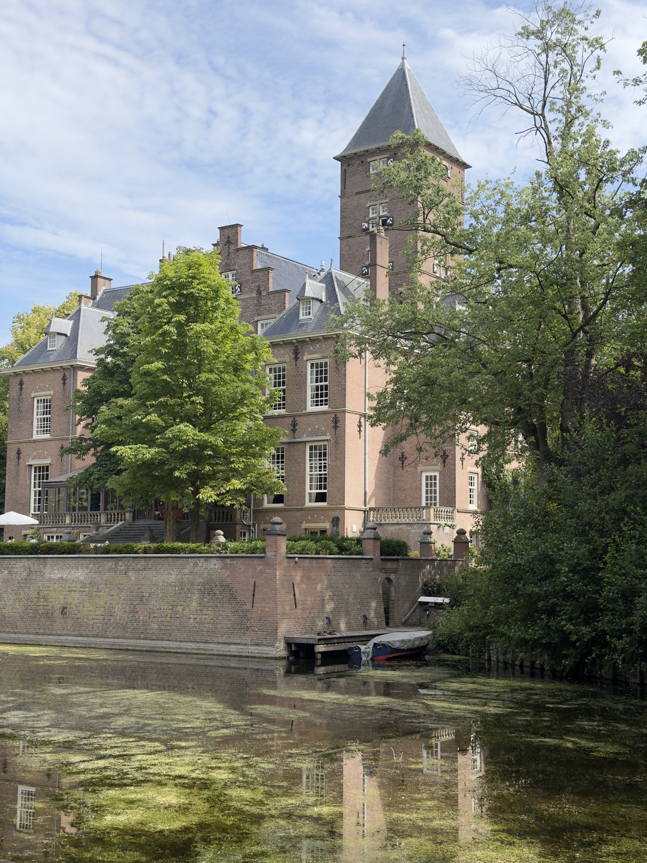 Brick castle with turret and steep roofs reflected in a green, algae-covered moat.