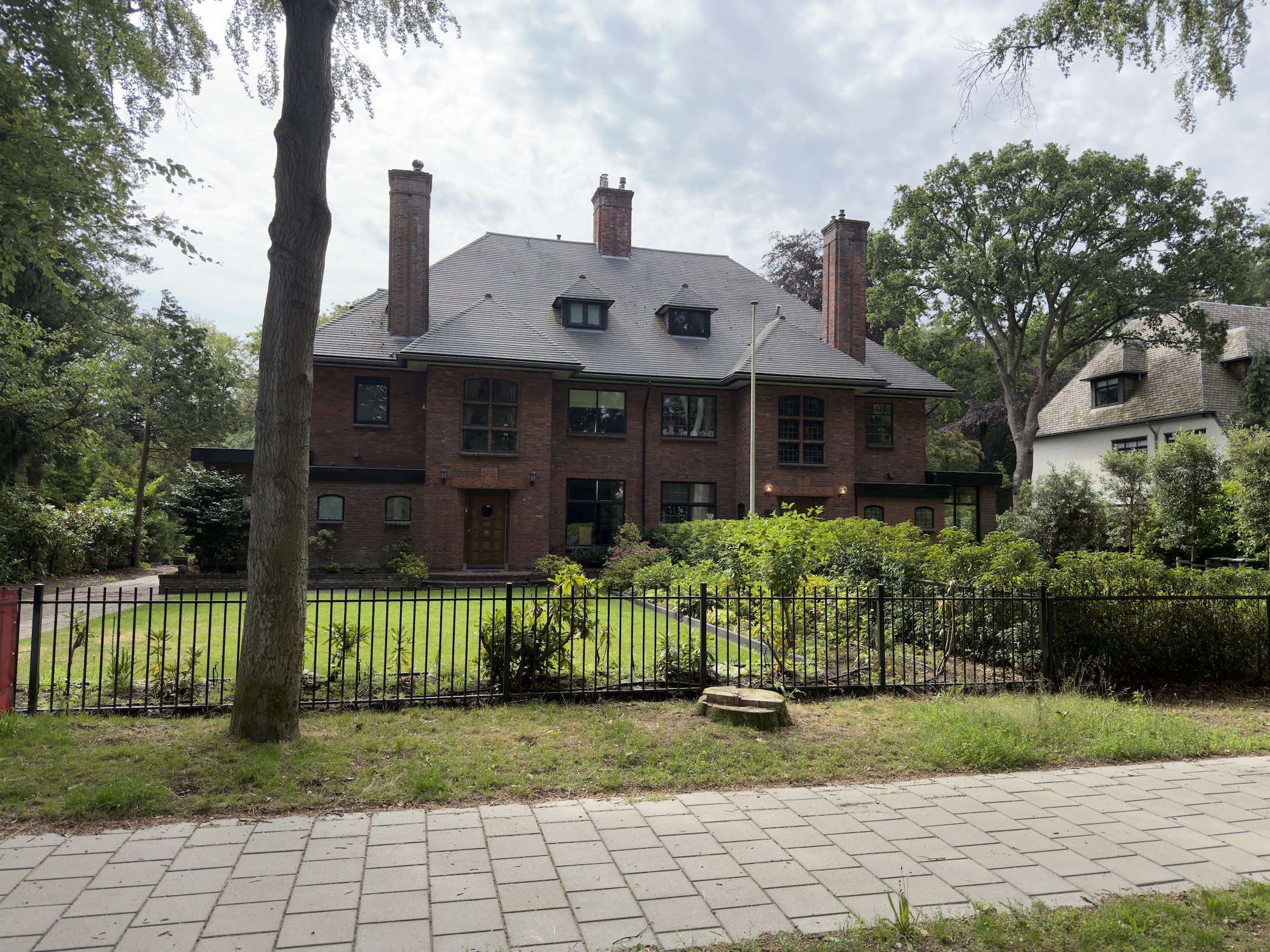 Large brick villa with tall chimneys behind a black iron fence and clipped garden.