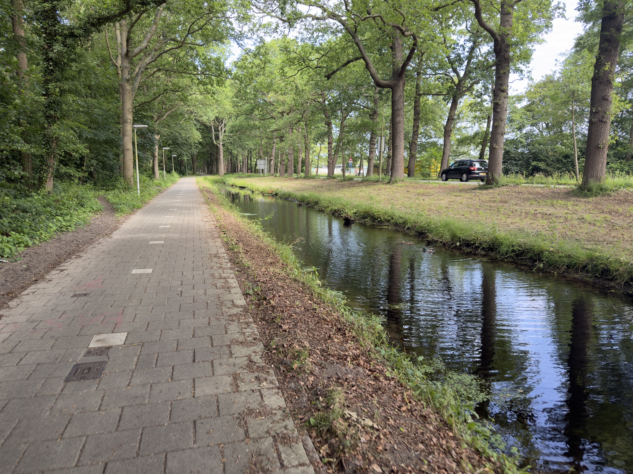 Brick-paved cycle path running alongside a narrow tree-shaded canal with a parked car.