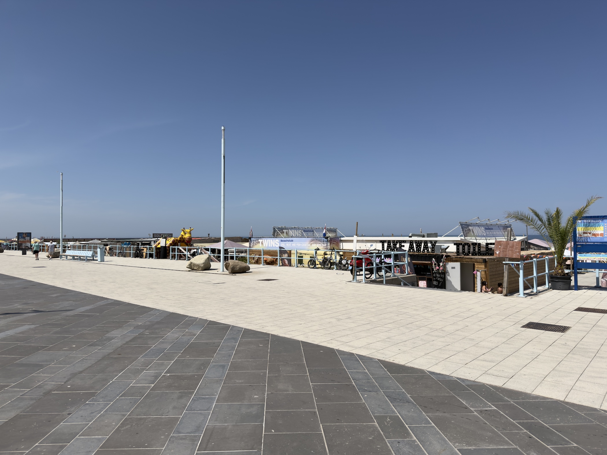 A wide tiled seafront promenade beside beach kiosks marked Take Away and Twins under blue sky.