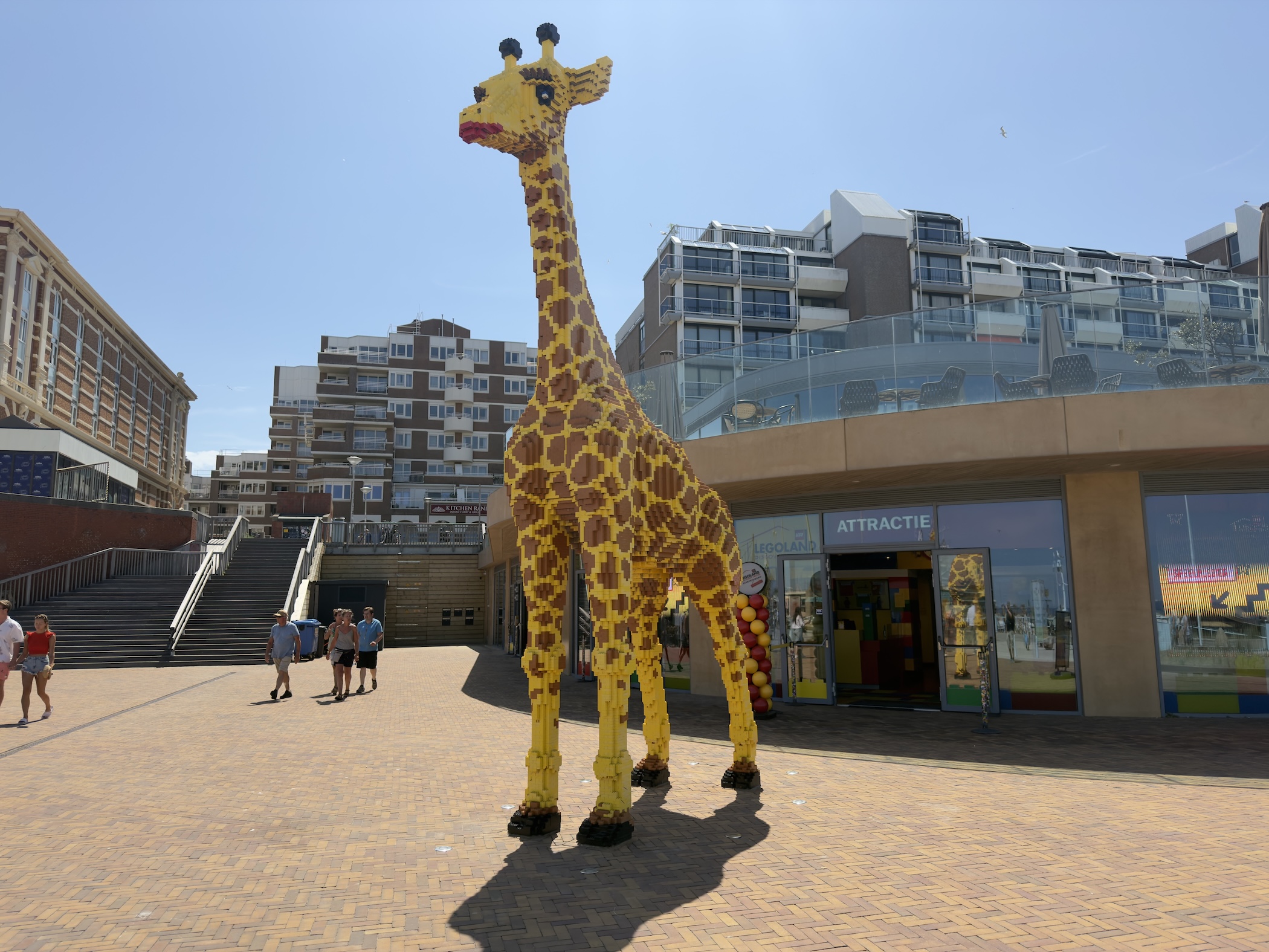 A giant yellow-and-brown giraffe sculpture towers over a paved square in front of apartment blocks.