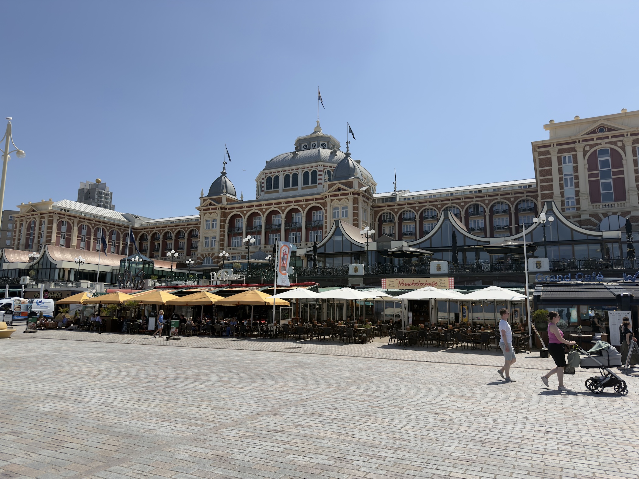 The grand domed Kurhaus building rises above seafront cafe terraces with yellow parasols.