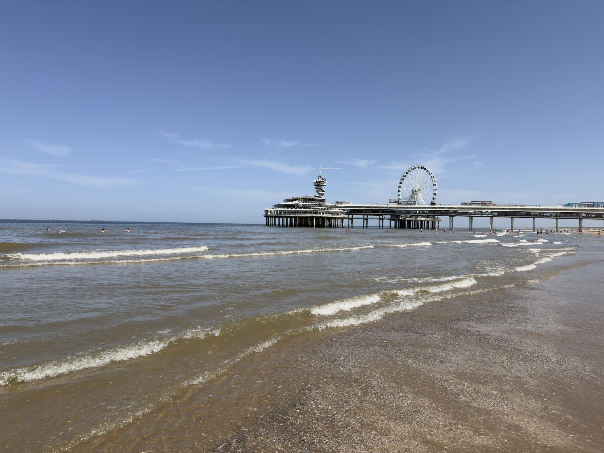 Gentle waves break on a sandy beach with Scheveningen pier and its Ferris wheel in the distance.