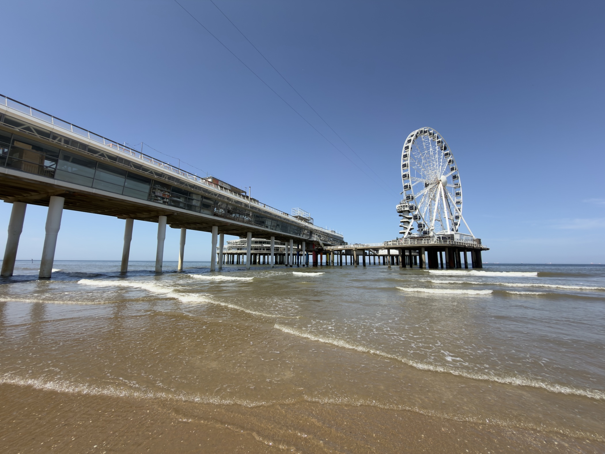 A low-angle view along Scheveningen pier on stilts with the white Ferris wheel under deep blue sky.