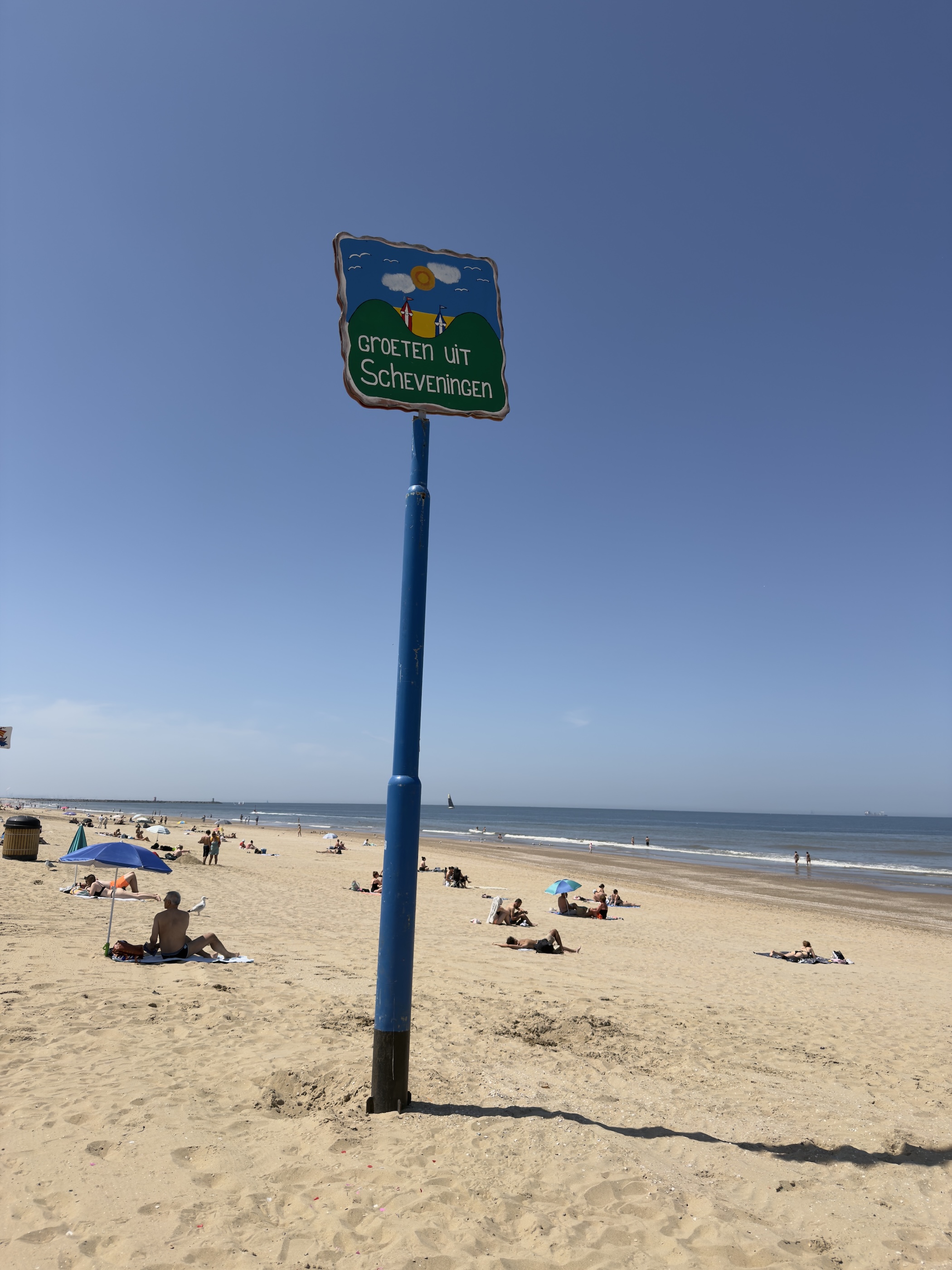 A blue post with a Groeten uit Scheveningen sign stands on a busy sandy beach by the sea.