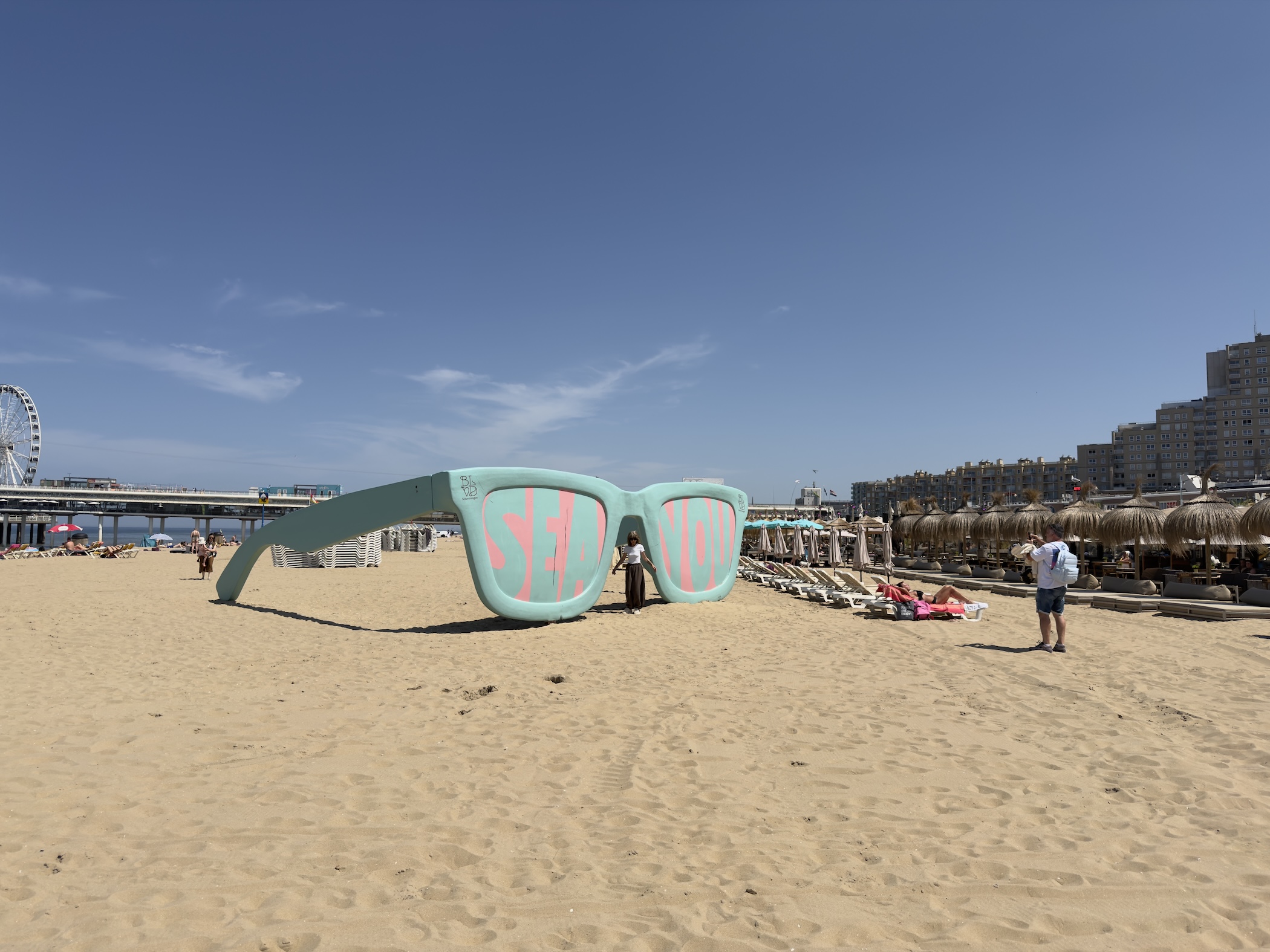 A giant pale green Sea You sunglasses sculpture sits on the beach near the pier and Ferris wheel.
