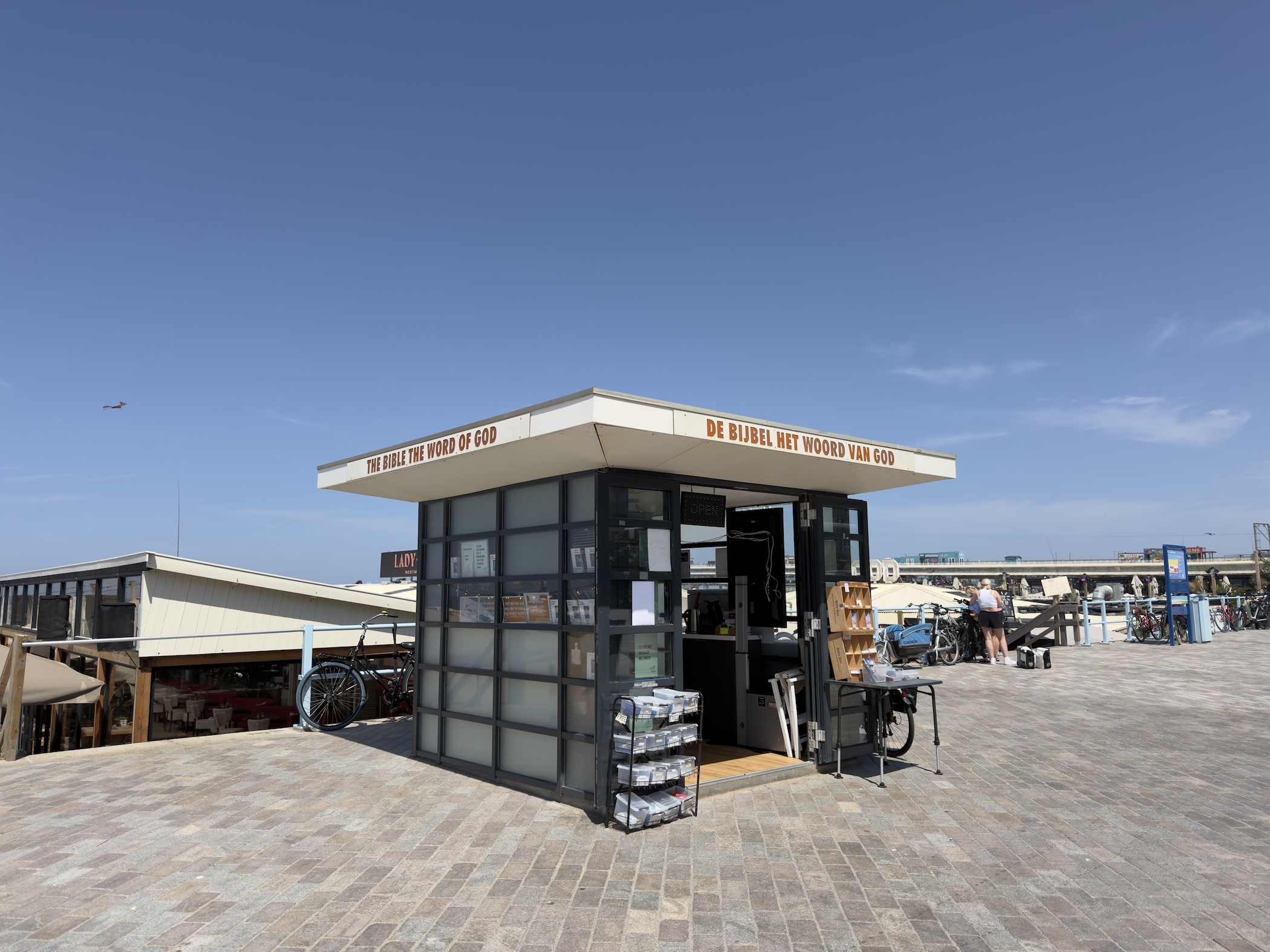 Glass-walled kiosk reading The Bible The Word of God on a sunny brick promenade with parked bikes.