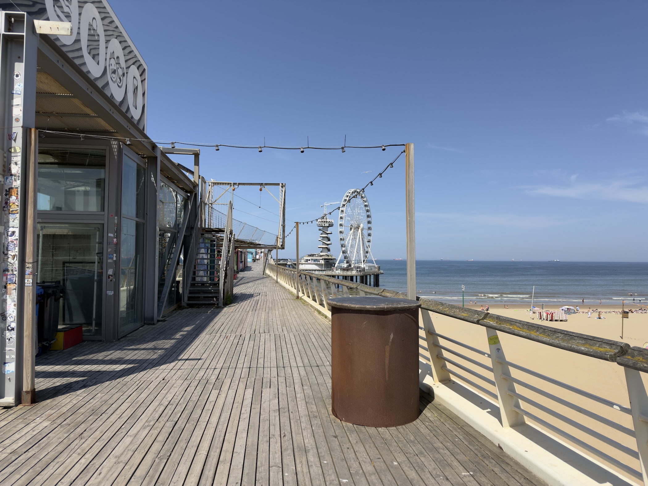 Wooden pier boardwalk leading towards a Ferris wheel, with the sandy beach and sea beyond.