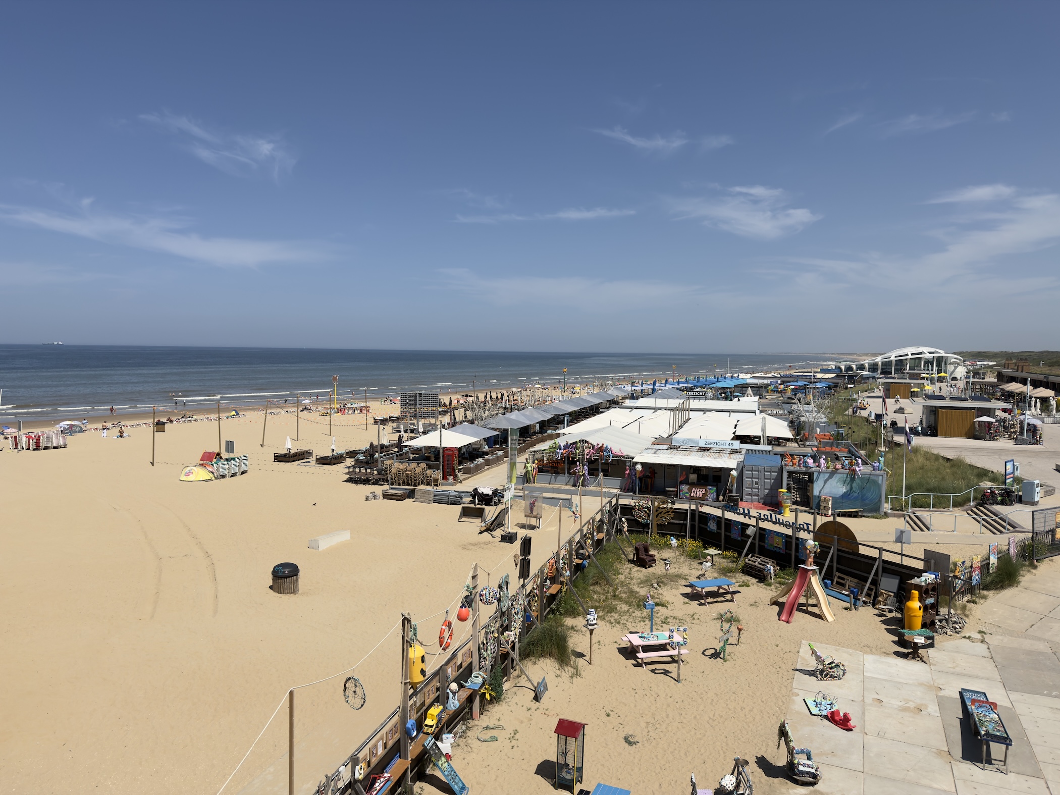 Elevated view over beach clubs, terraces and a playground on the wide sandy shore under blue sky.