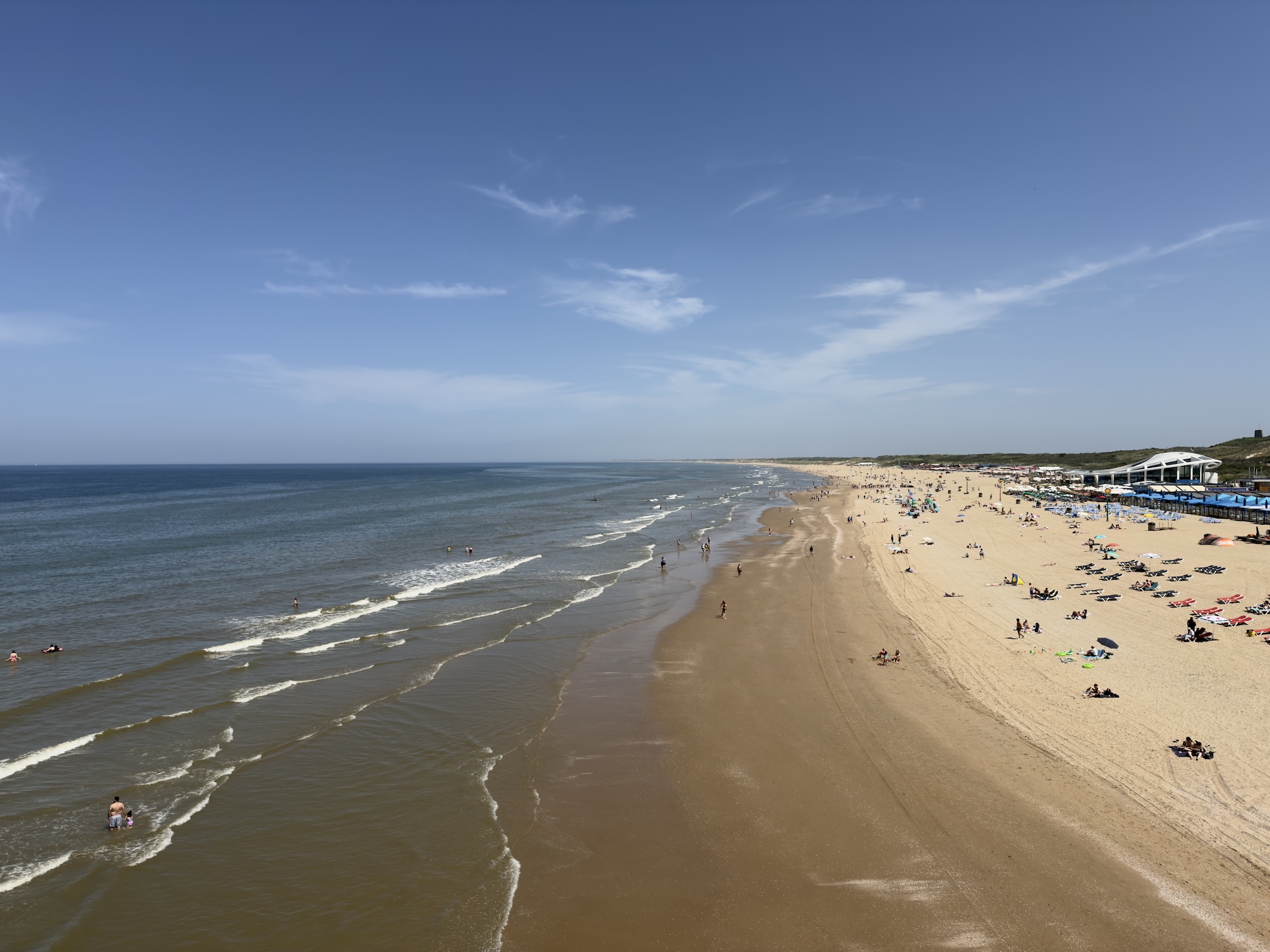 Aerial view of breaking waves rolling onto a curving beach dotted with sunbathers and loungers.