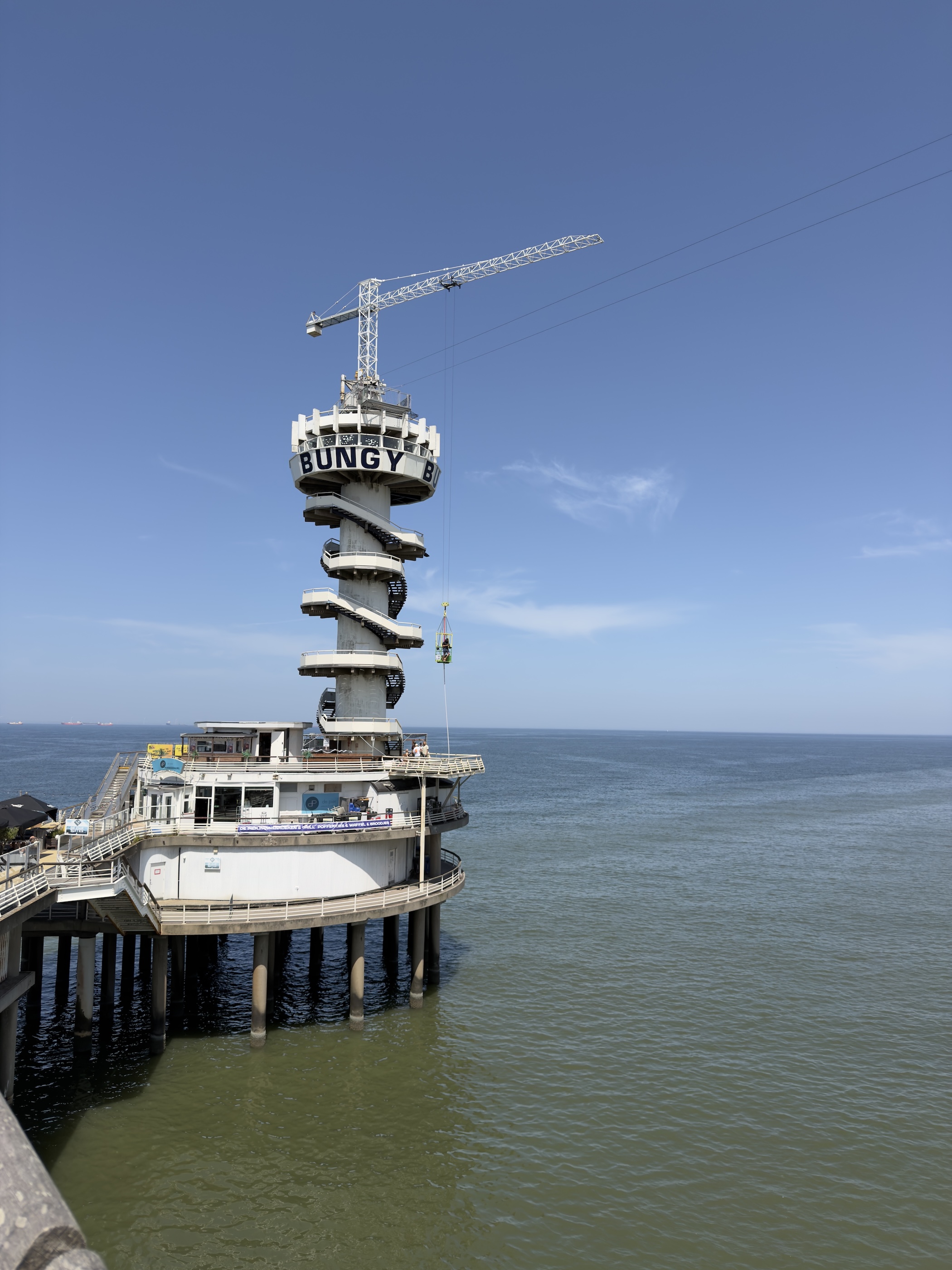Spiral bungy jump tower with a crane rising above a round pier building over the green sea.