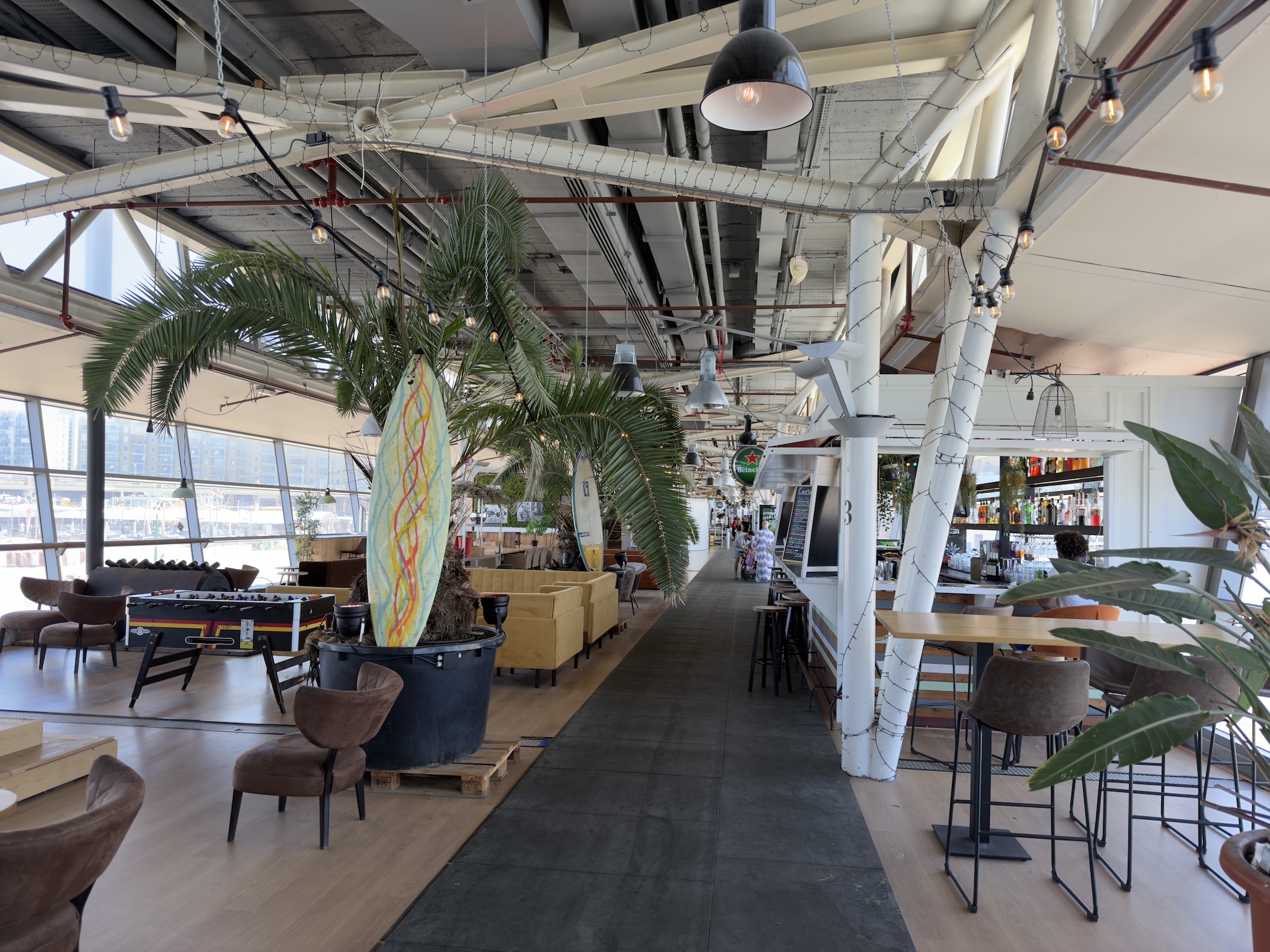 Airy beach club interior with palm trees, pendant lights, bar stools and a hanging surfboard.