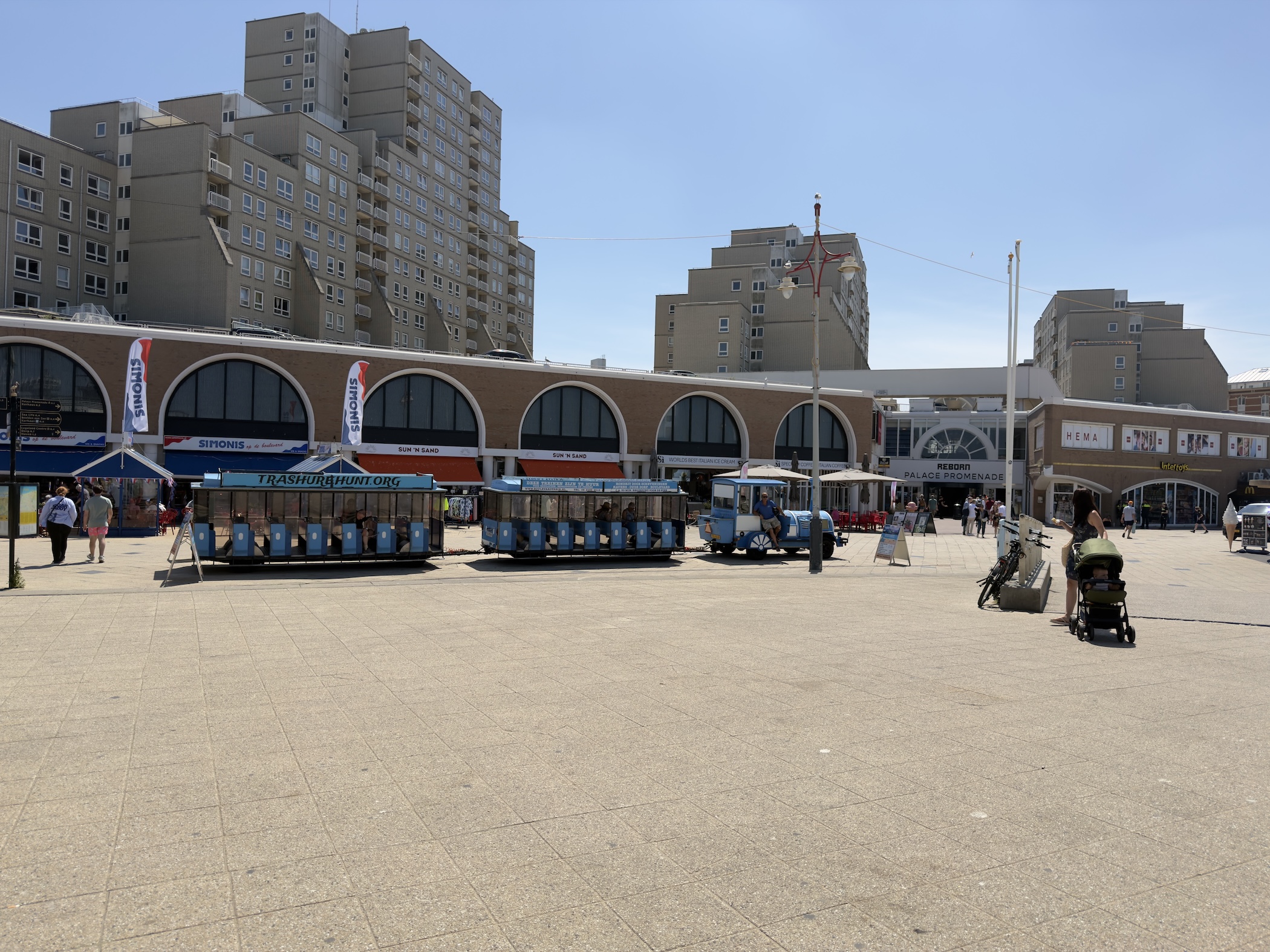 Blue tourist road train parked on a tiled square before arched shops and tall apartment blocks.