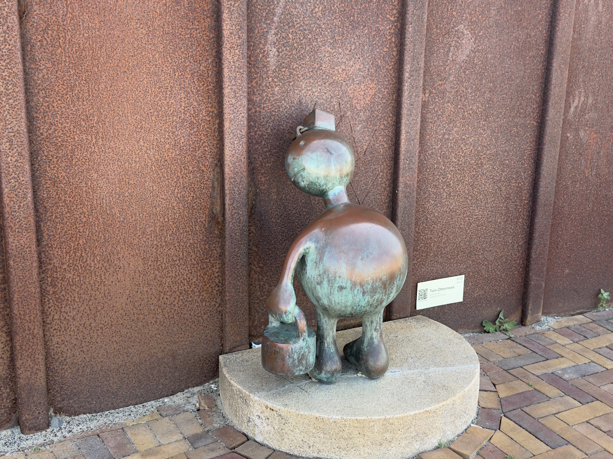 Bronze cartoon-style cat sculpture against a rusted steel wall, set on paved stone.