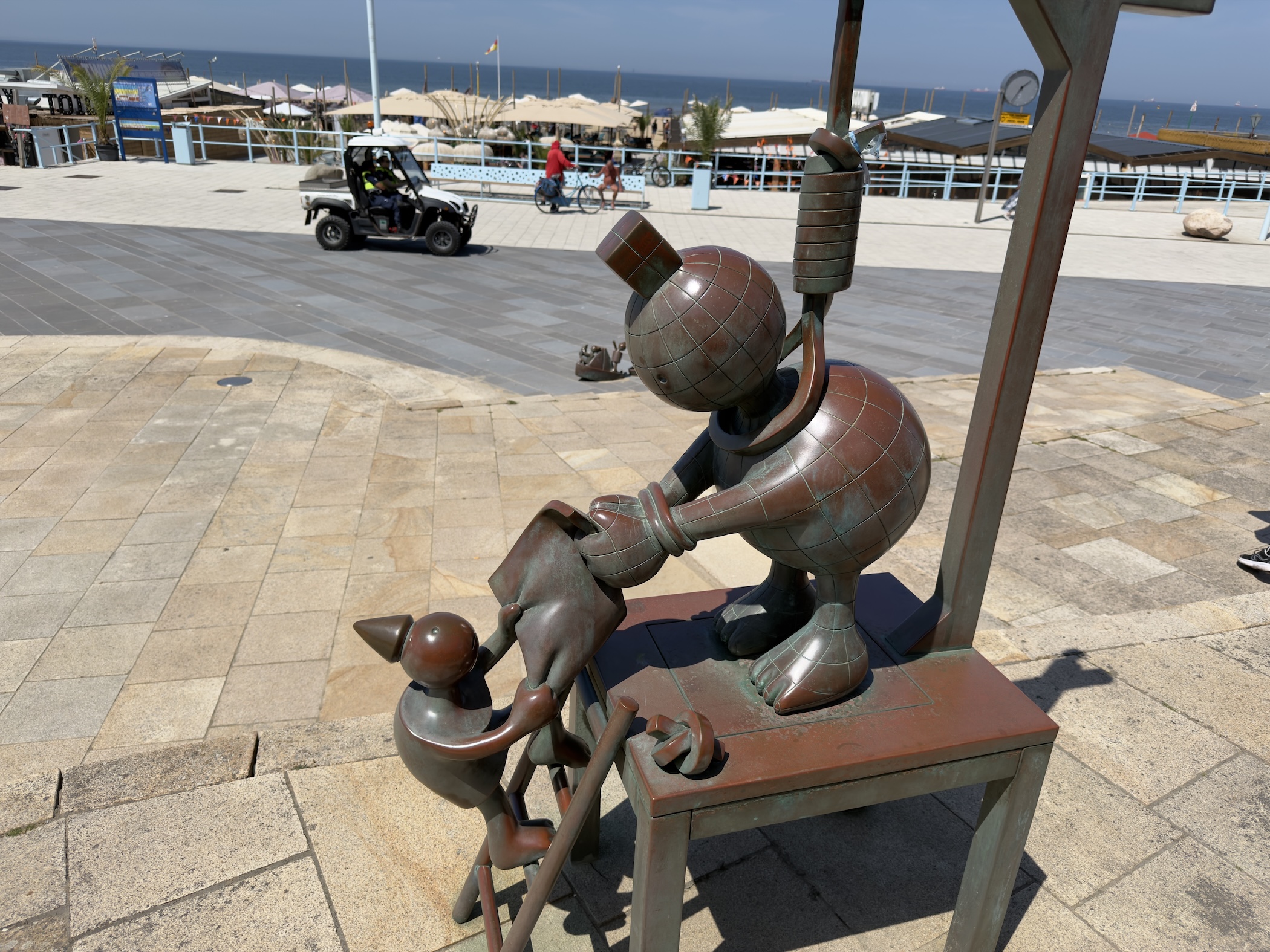 Whimsical bronze figures at a desk on a sunlit seafront promenade, sea beyond.