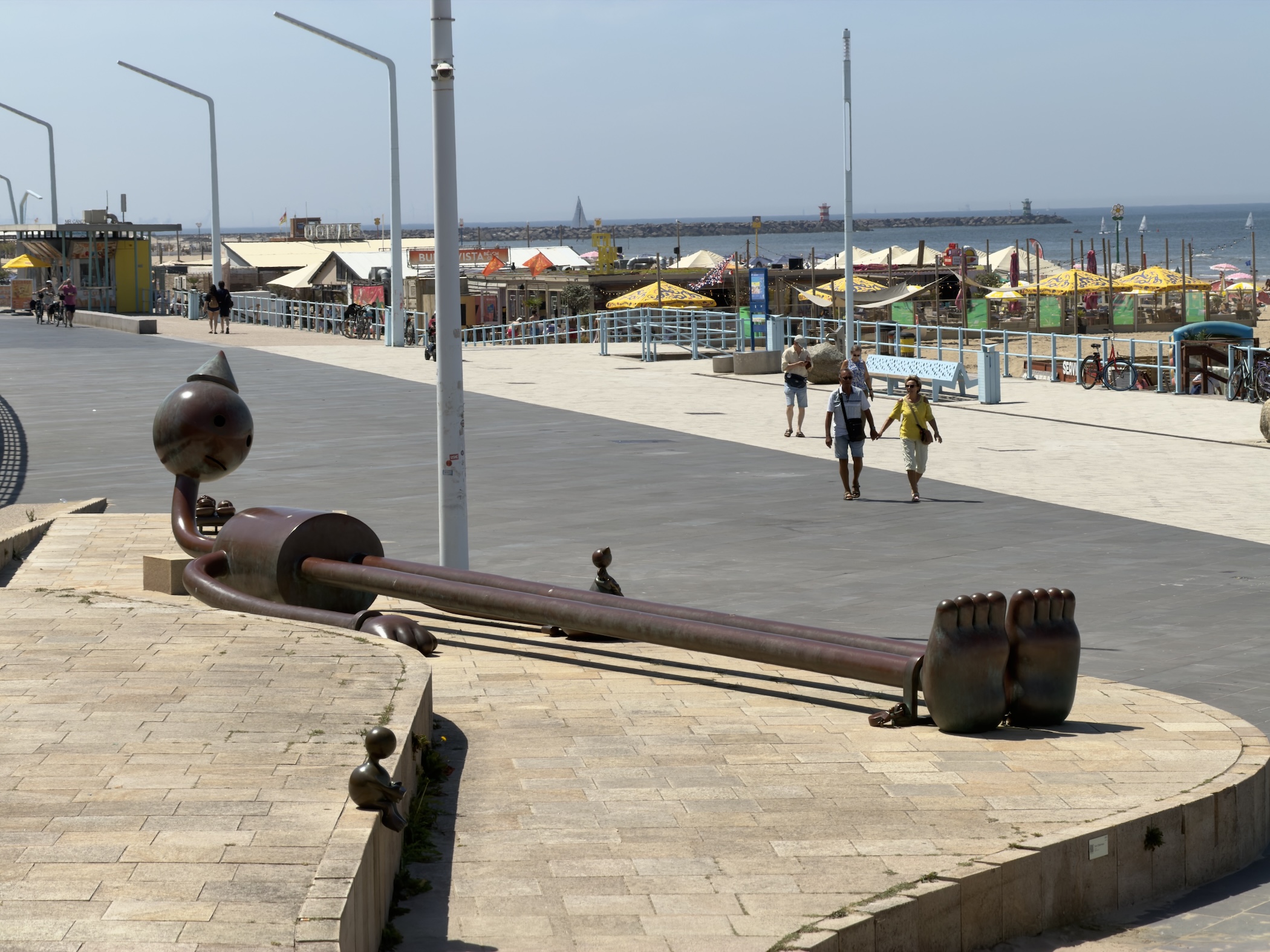 Long reclining bronze sculpture on the promenade as people walk past beach huts behind.