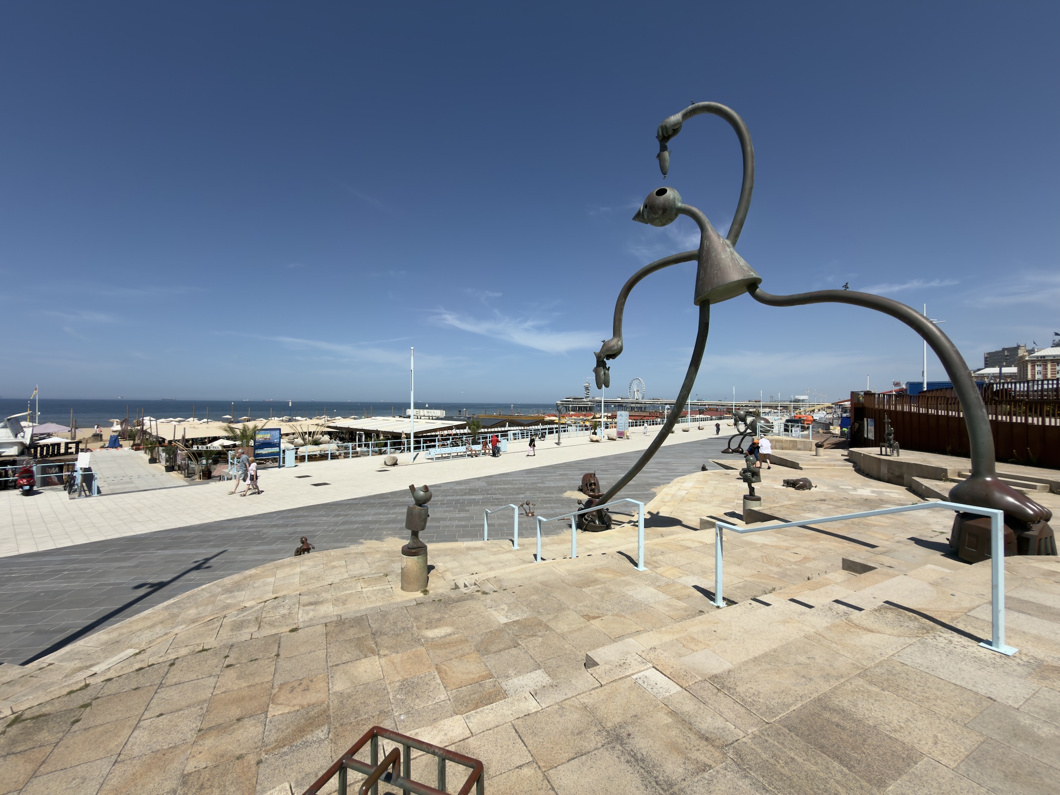 Tall curling tendril-like bronze sculpture arching over paved steps above a sunlit seafront.