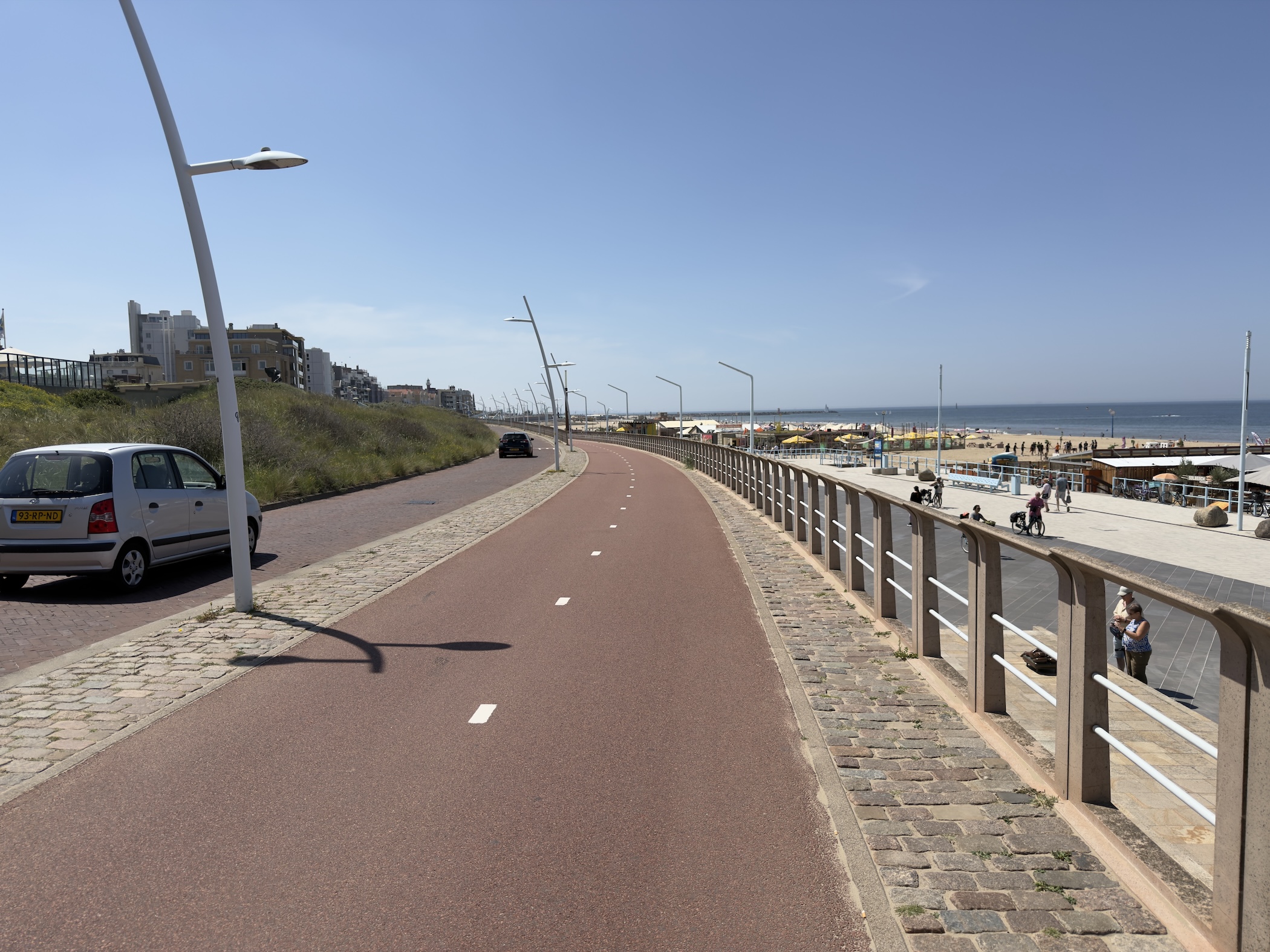 Empty red cycle path curving along the seafront beside a beach, parked car on the left.