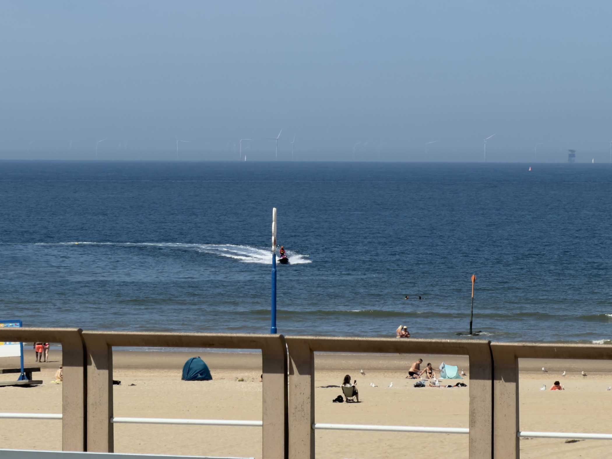 Sandy beach and deep blue sea with a jet ski, swimmers and distant offshore wind turbines.