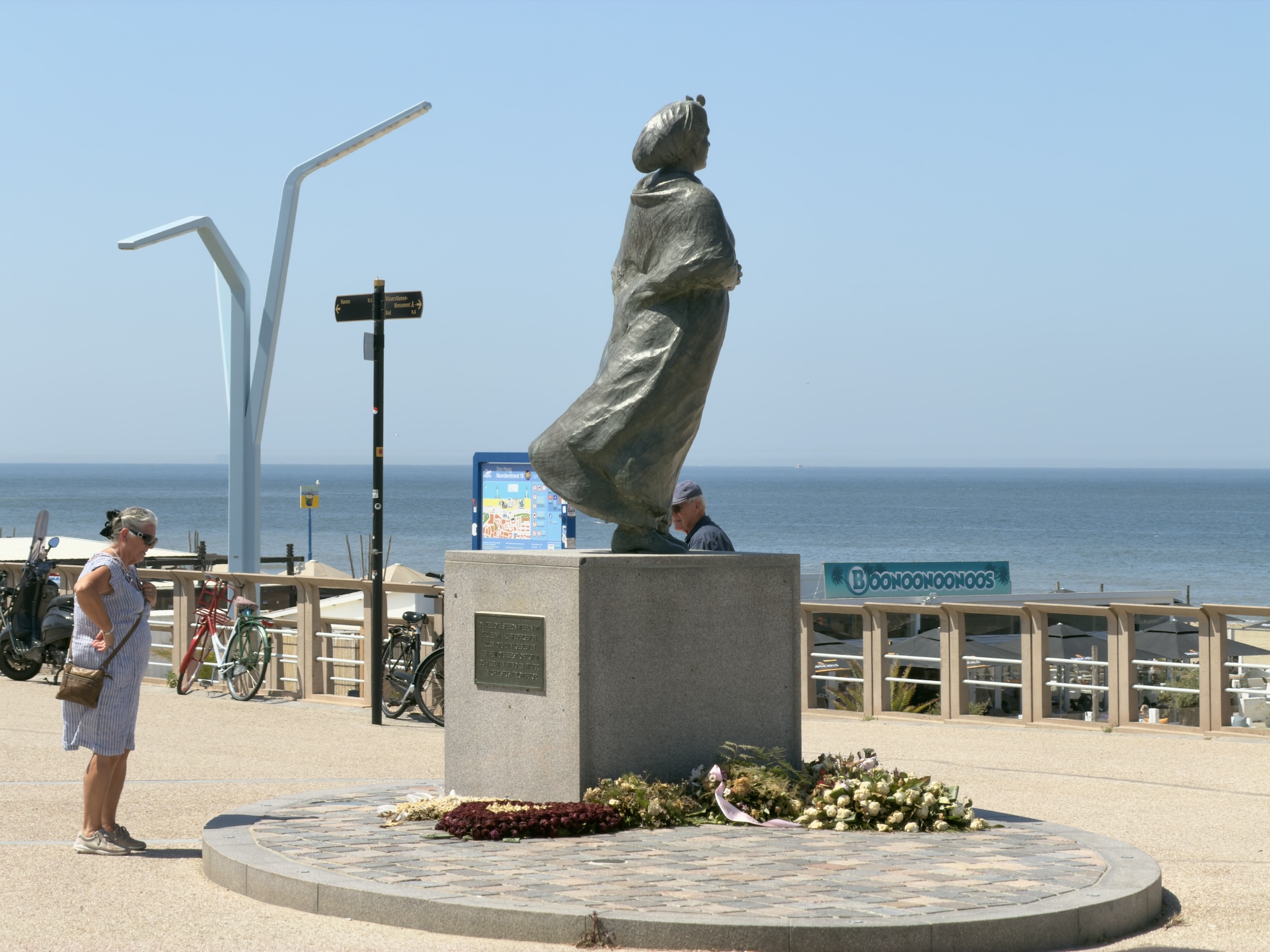 Bronze statue of a robed woman on a plinth with flowers laid below, bicycles and sea behind.