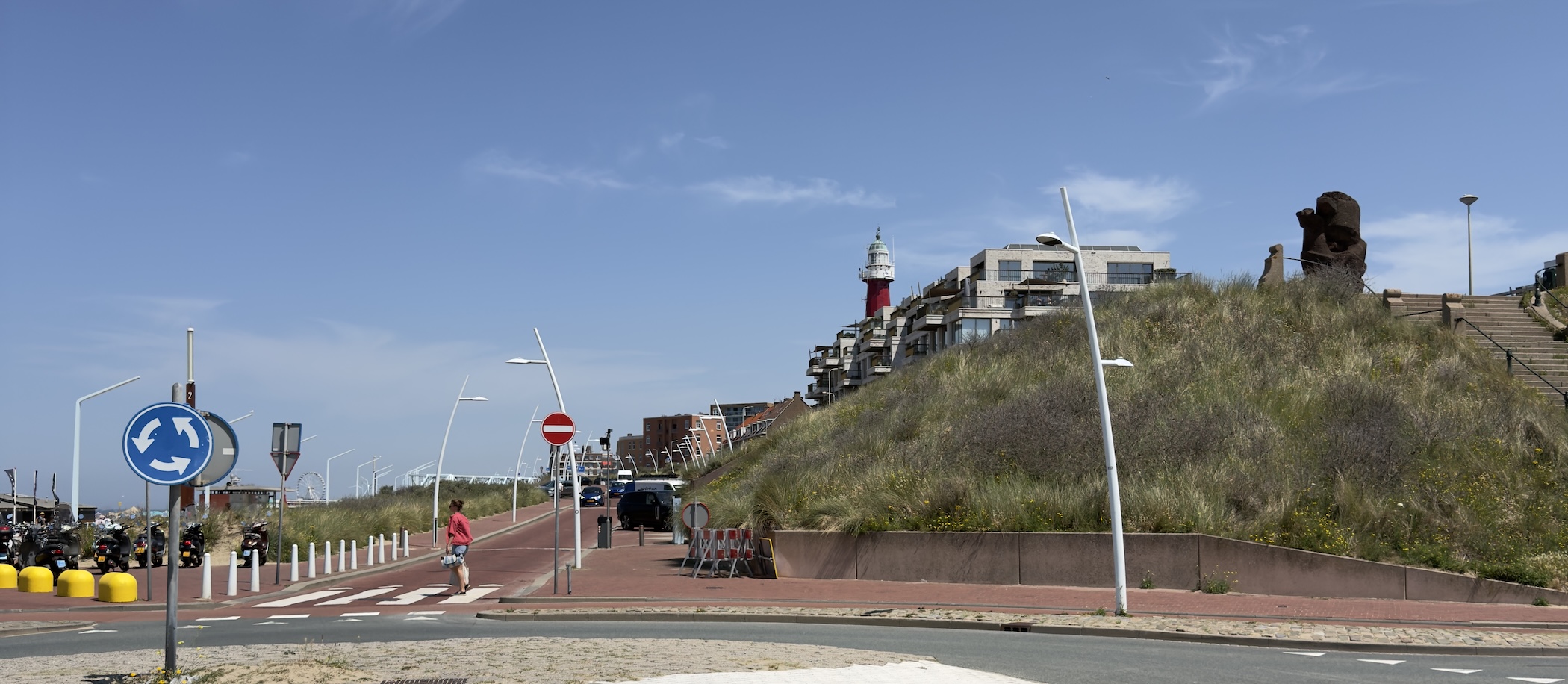 Lighthouse and apartments rising above a grassy dune beside a cyclist on a red bike path.