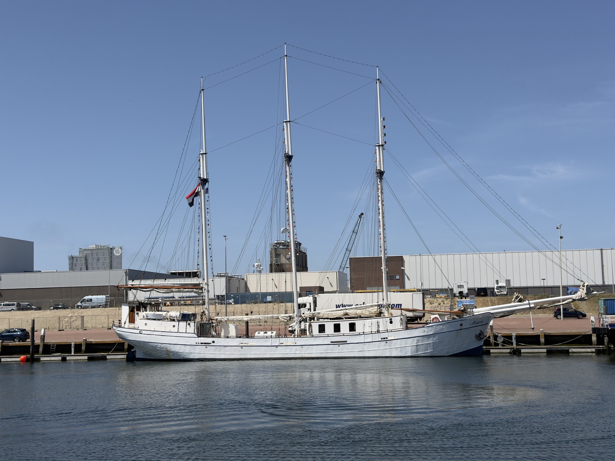 A three-masted white sailing ship berthed in calm dock water before industrial sheds.