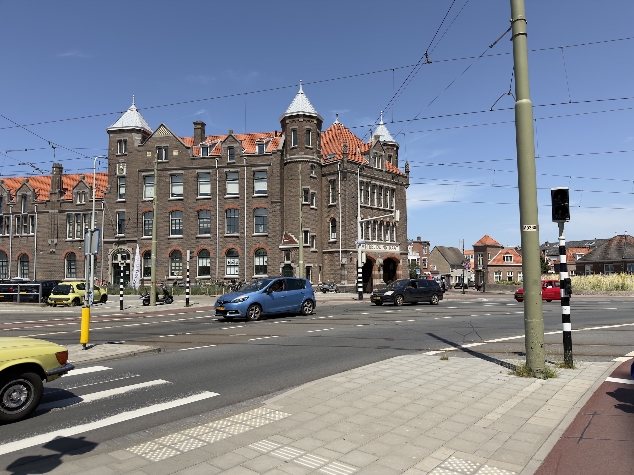A turreted brown brick building beside a tram-wire crossroads under a clear blue sky.