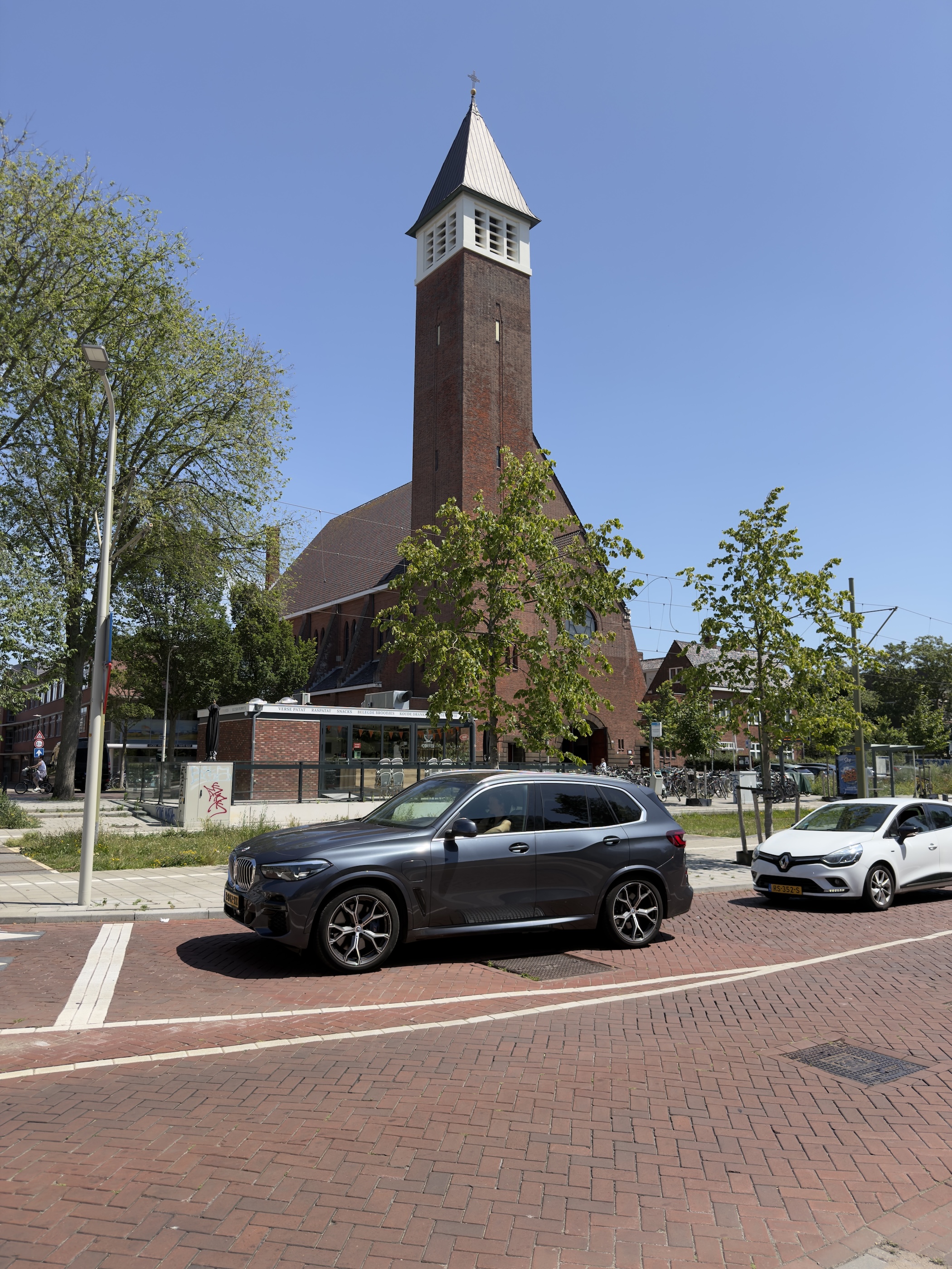 A tall brick church with a white spire rising behind parked cars on a paved square.