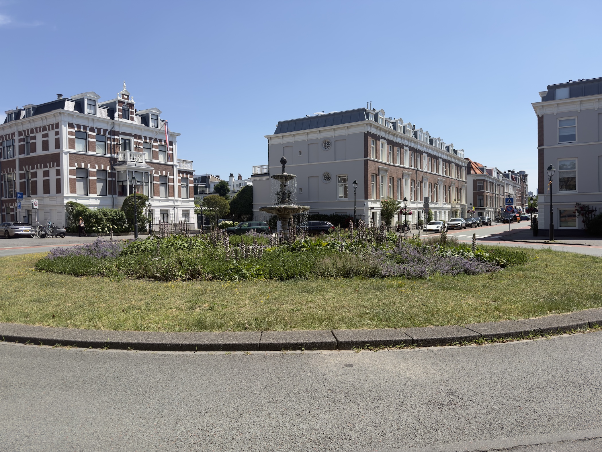 An ornamental fountain and flower bed on a grassy roundabout before stately townhouses.