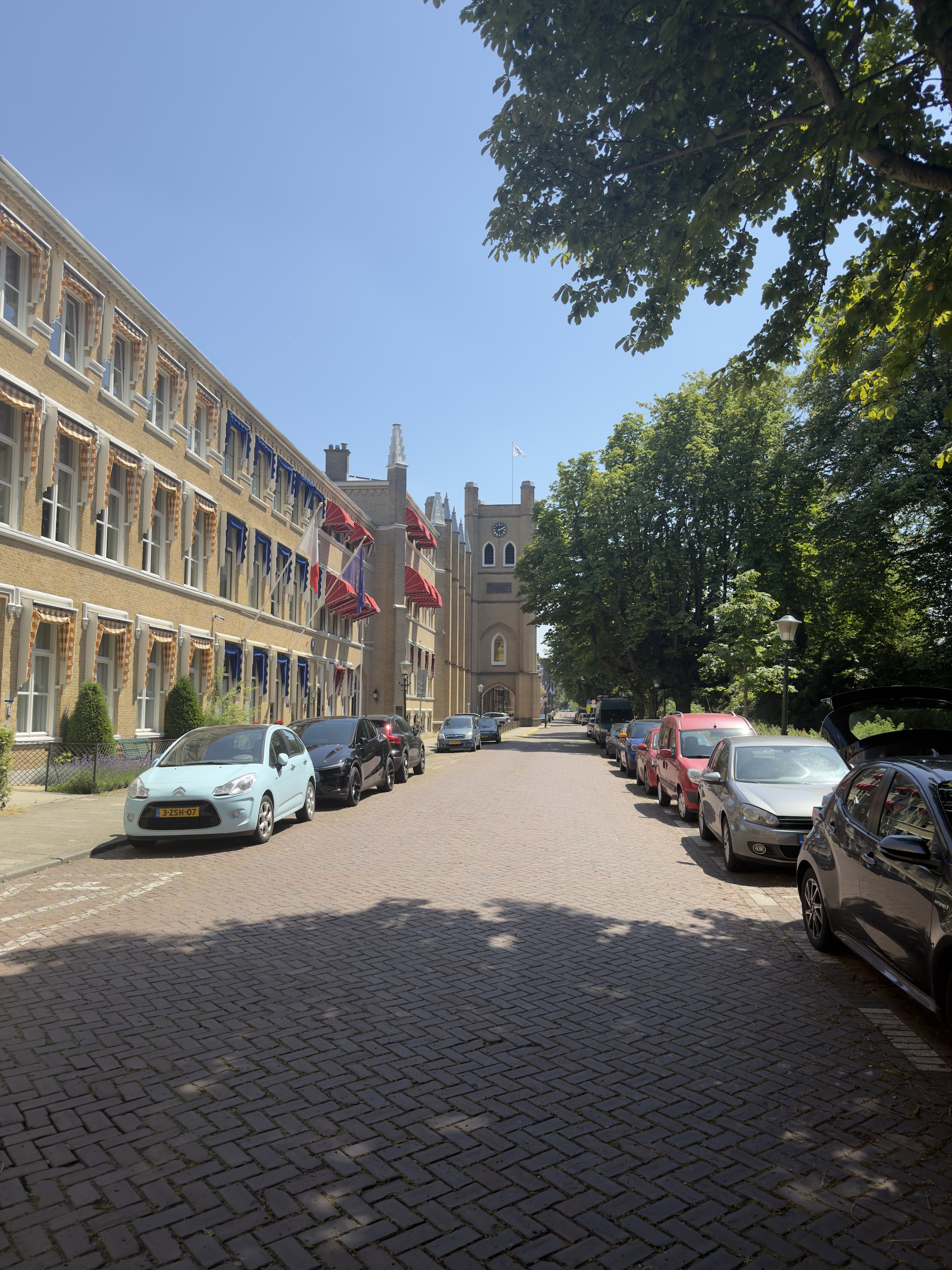 A cobbled residential street with parked cars, orange shutters and a chapel at its end.