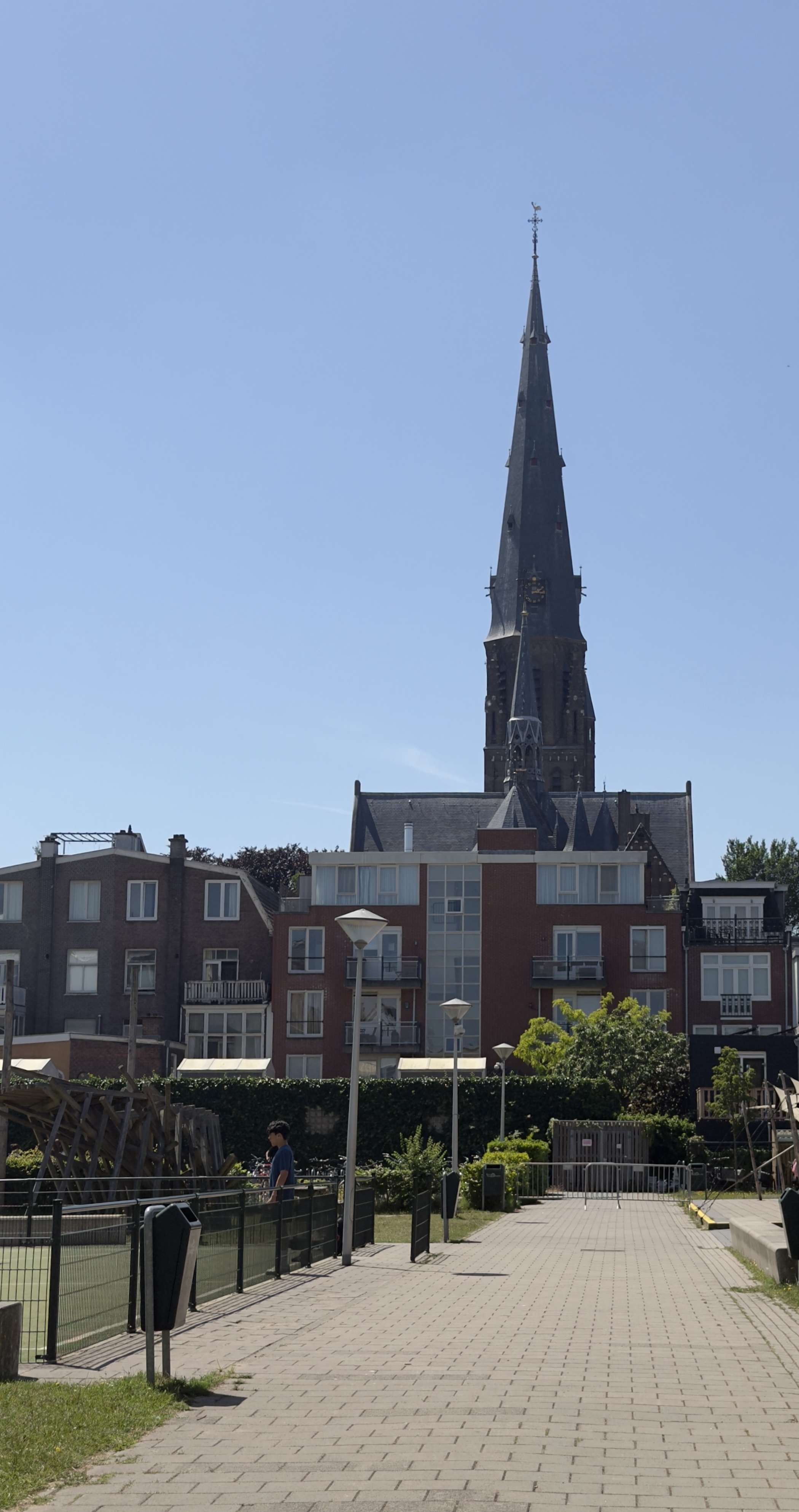A dark grey church spire towering over red brick flats beside a paved playground walkway.