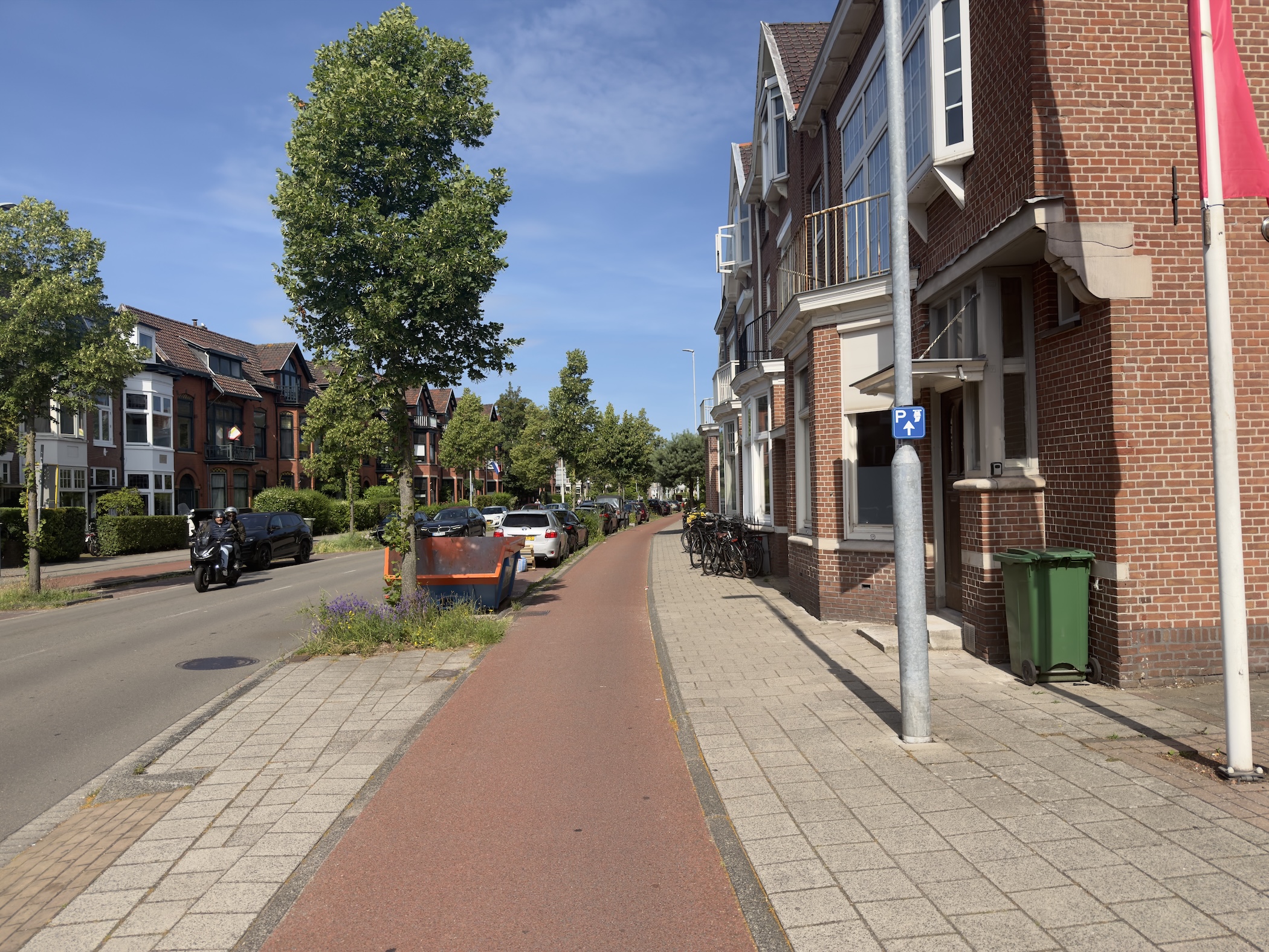 A red-paved cycle path running between brick houses and parked cars on a quiet sunny street.