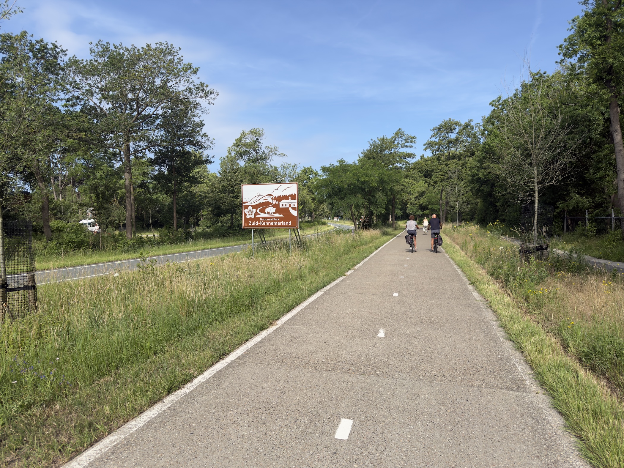 A wide cycle path crossing grassland towards the Zuid-Kennemerland park entrance sign, two cyclists ahead.