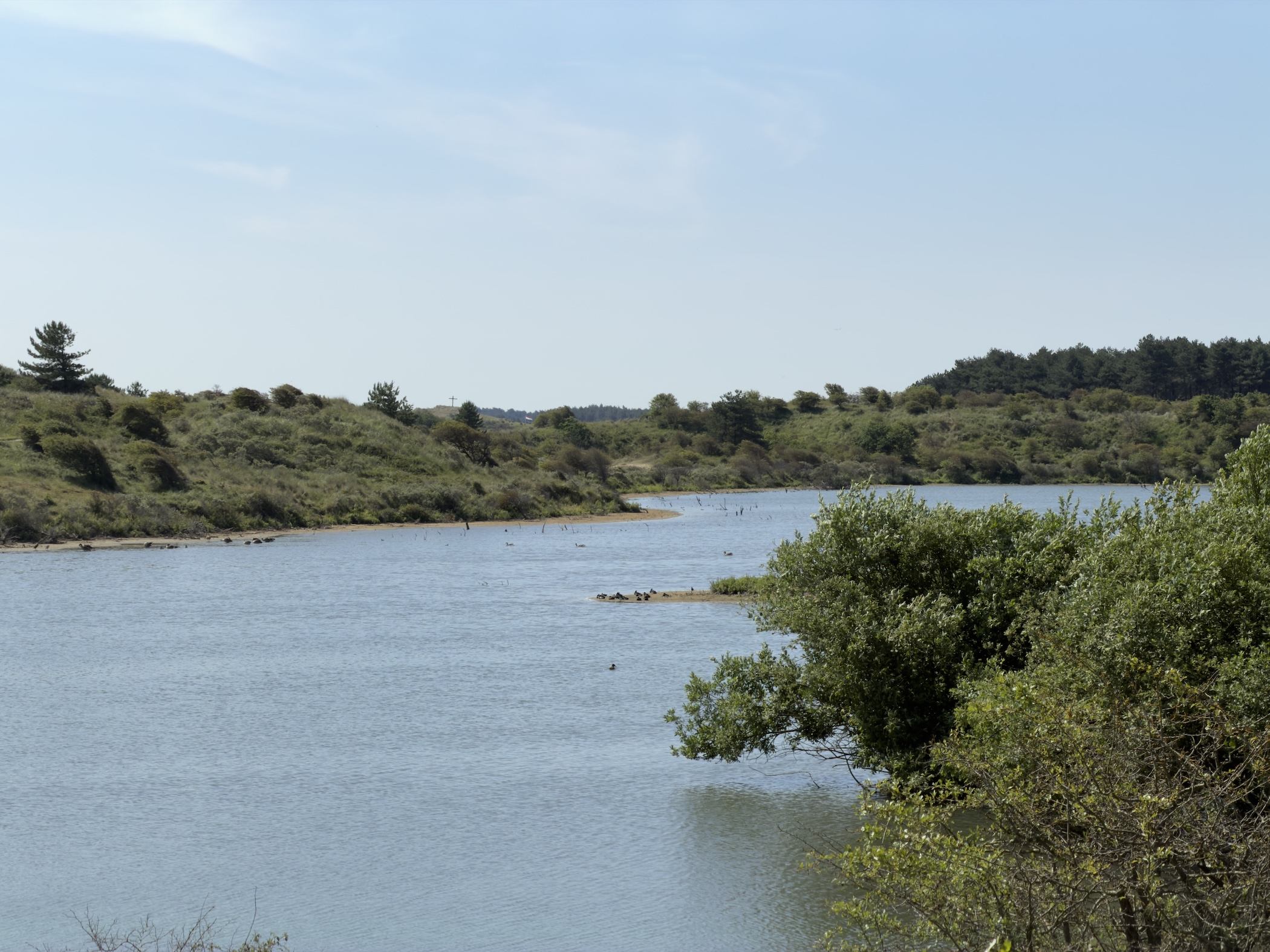 A calm dune lake fringed by green shrubs and low wooded slopes.