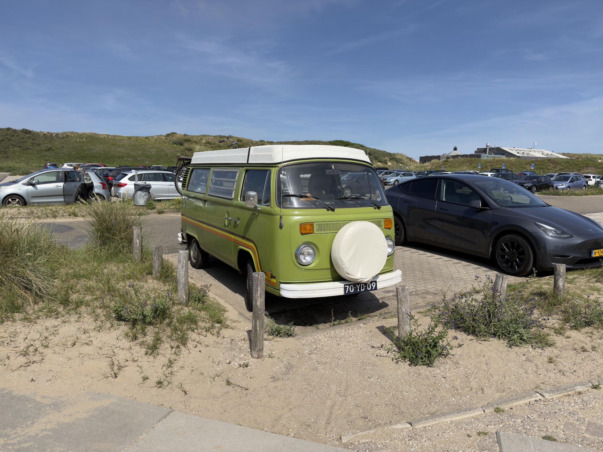 A green vintage Volkswagen camper van parked on sand beside a Tesla in a dune car park.