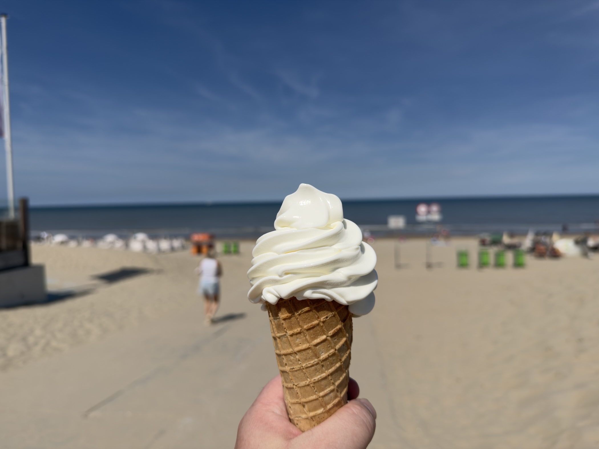 A hand holding a soft vanilla ice cream cone above a sunlit sandy beach and sea.