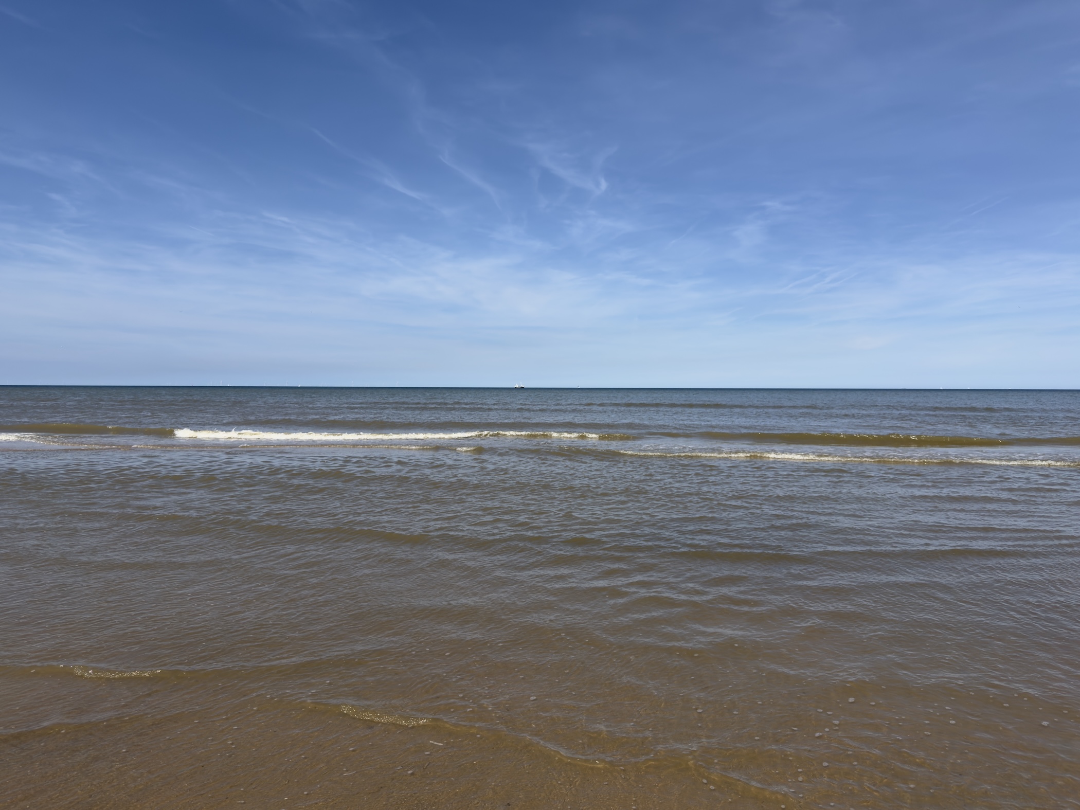 Gentle brown breakers rolling onto a flat sandy shore beneath a hazy blue sky.