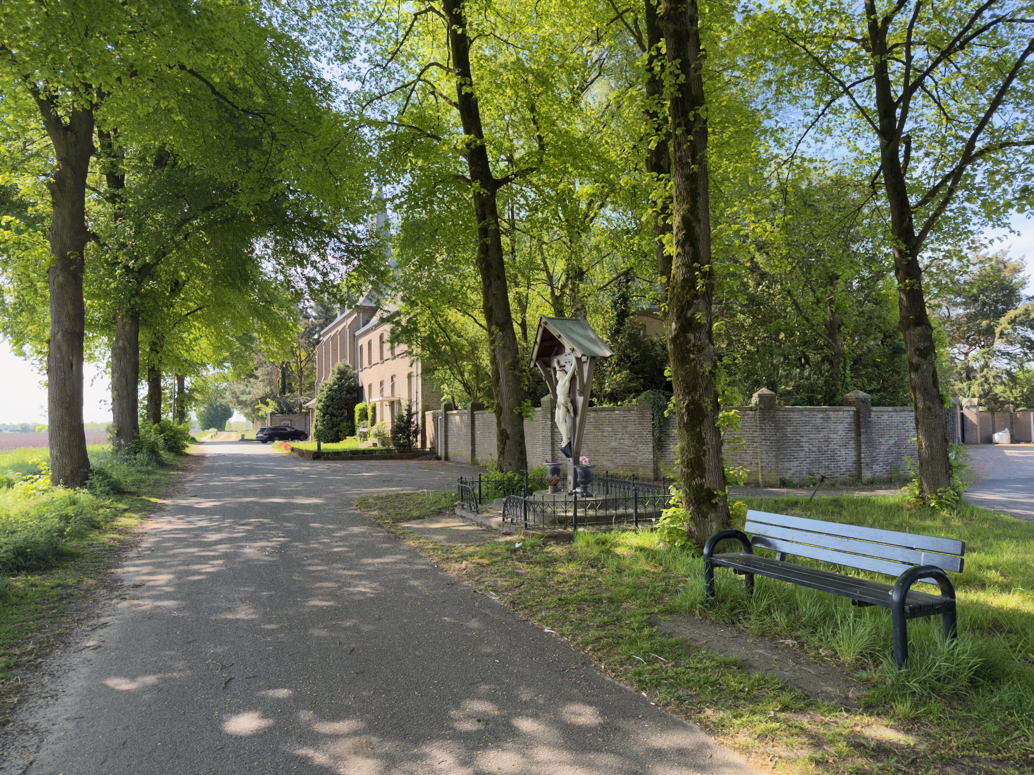 Tree-lined lane with a green bench beside a small wayside shrine and crucifix