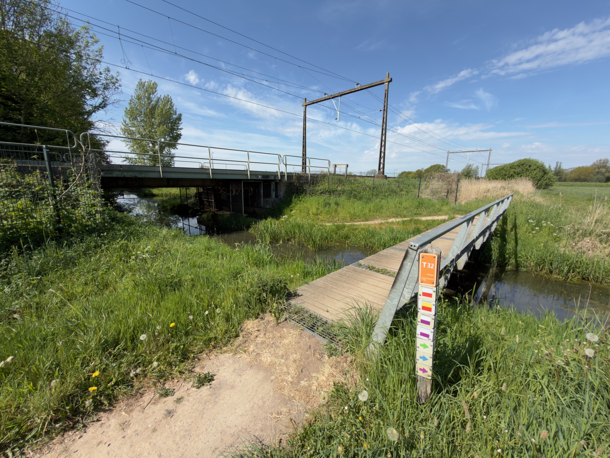 Wooden footbridge crossing a small stream beside a railway bridge with overhead wires
