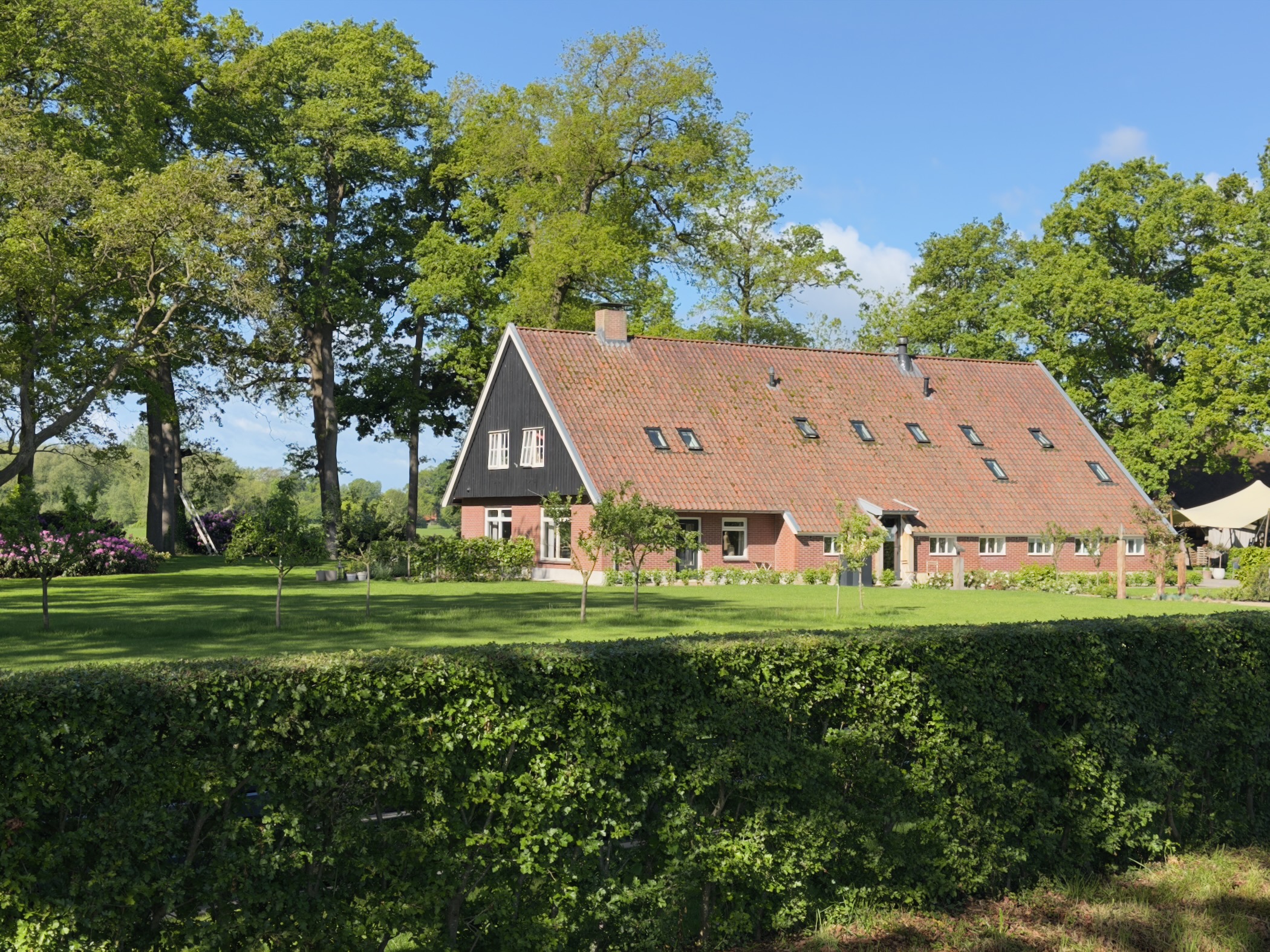 Brick farmhouse with red-tiled roof set behind a clipped hedge among tall trees