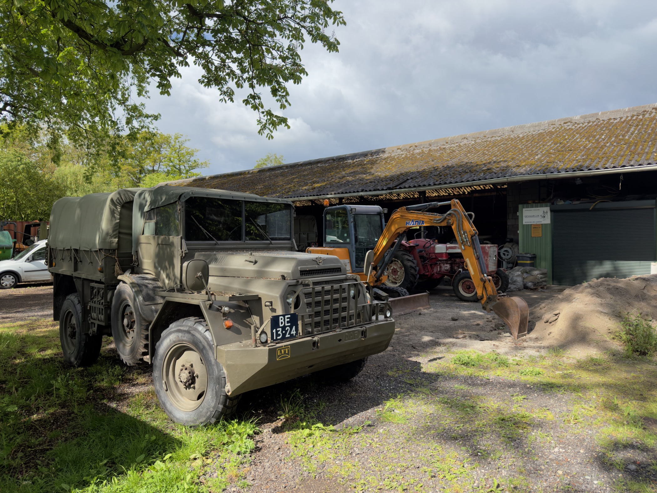 Olive-green ex-military truck parked beside a barn with an orange excavator at work