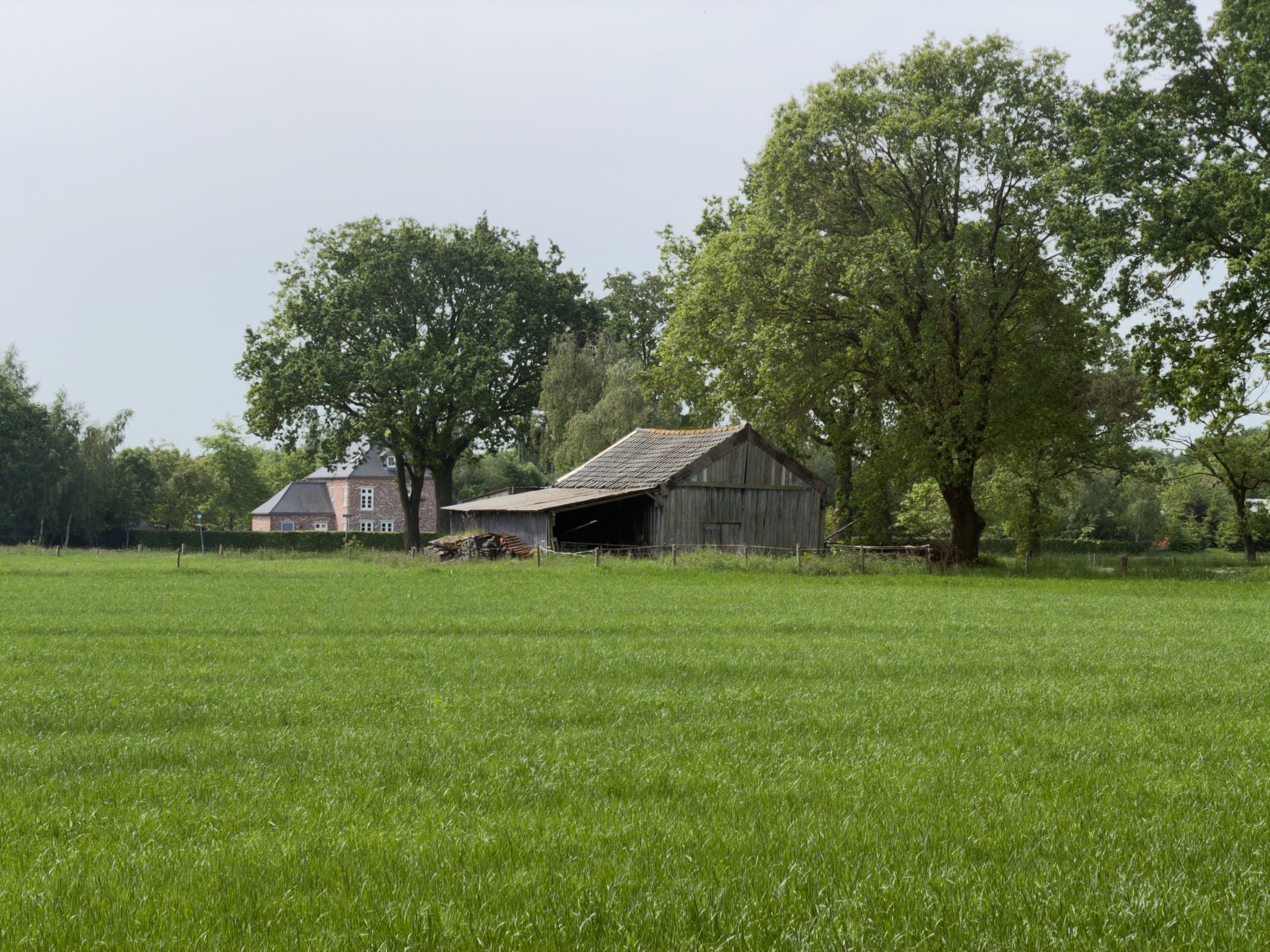 Old wooden hay barn behind a fresh green meadow with a farmhouse to the left