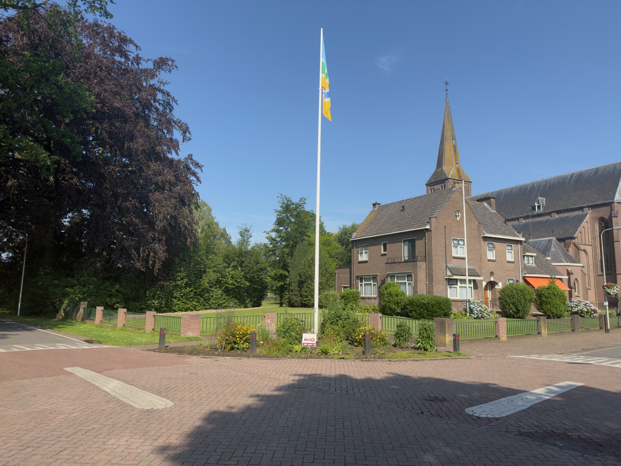 A village square with a flagpole flying the Overijssel flag, beside a brick church with a slender spire and houses behind a hedge.