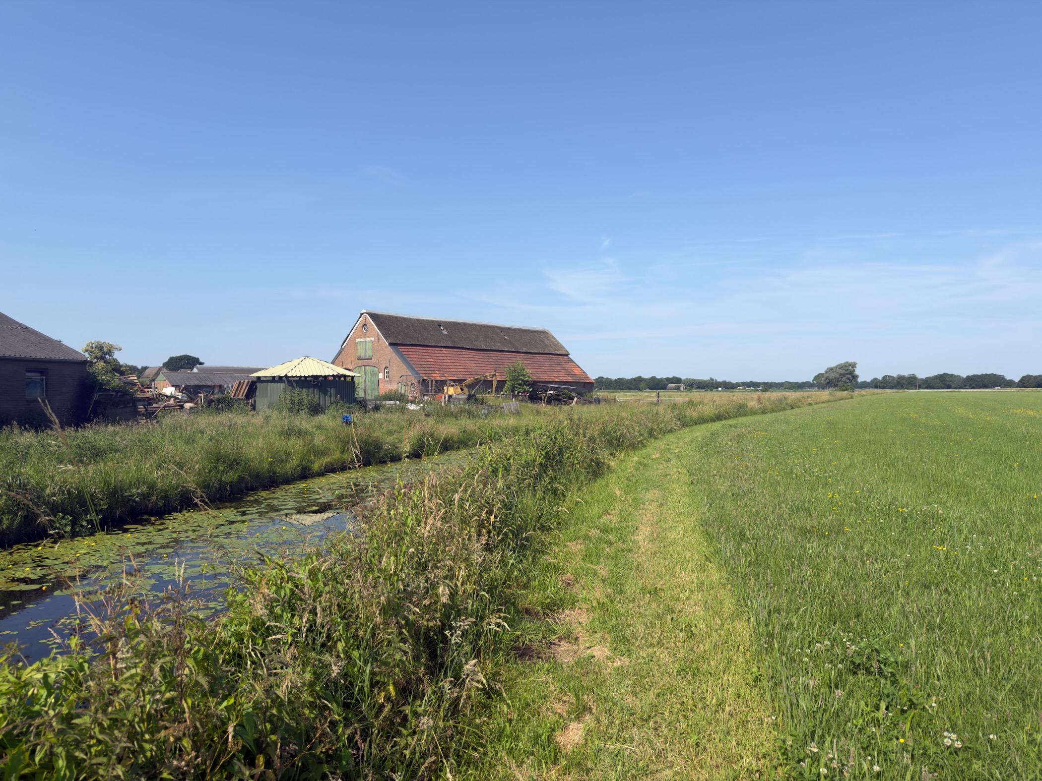 A grass path running along a ditch beside a brick farmhouse, with green fields stretching to a distant tree line.