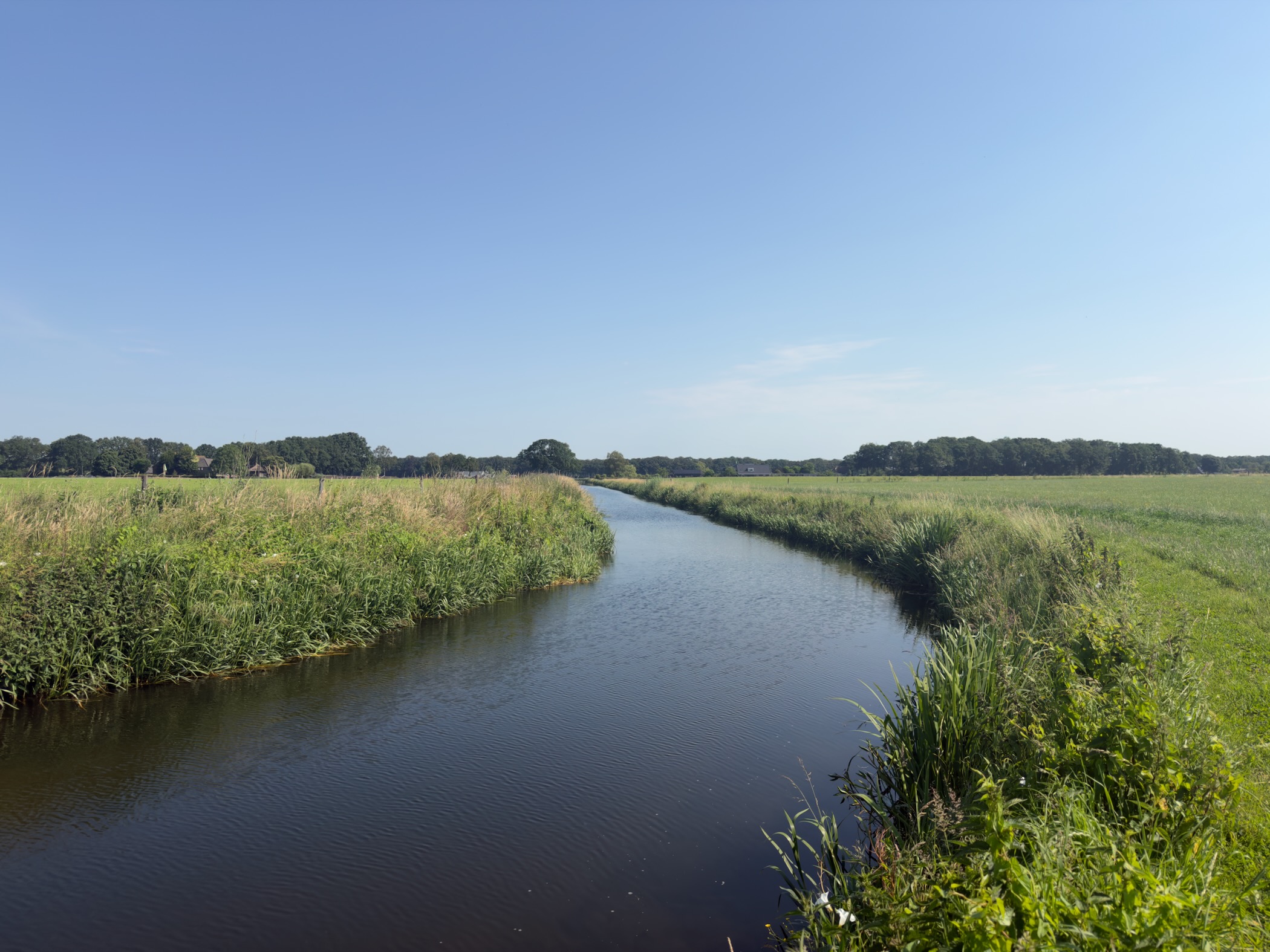 A calm watercourse winding between grassy banks across flat farmland towards a distant tree line.