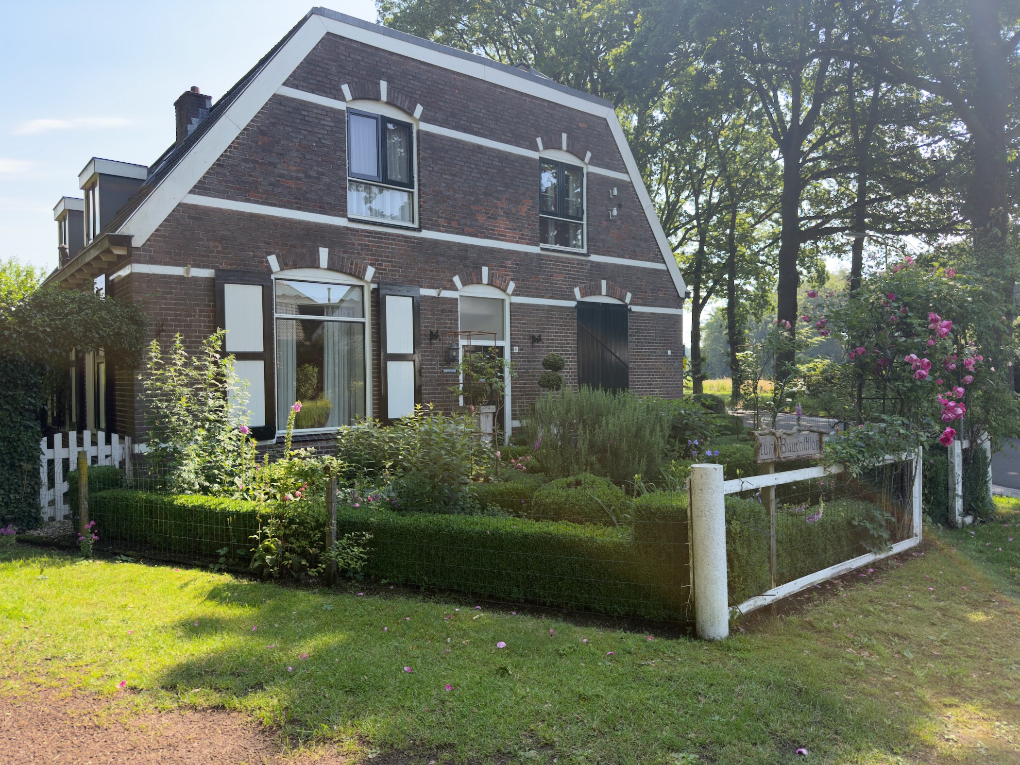 A brick house with white shutters and a gambrel roof, fronted by a low hedge and flowering roses.